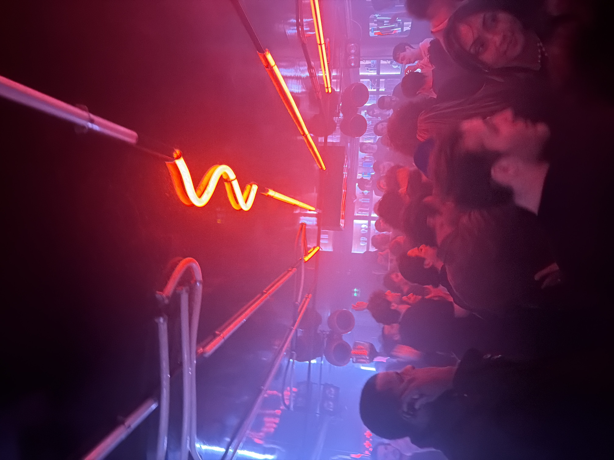 The image shows a dimly lit interior space filled with people, likely in Paris, France. The perspective is tilted, giving an unusual angle.
People: A crowd of people is present, mostly faces and heads visible, suggesting a tight space. Their expressions are not clear due to the low lighting and angles. Their actions are unclear, but the density suggests they might be observing something or participating in an event.
Objects: The room is illuminated by neon lights. Straight neon tubes are aligned horizontally, and one is bent into a zig-zag shape. Metal scaffolding or bars are visible in the lower-left corner, possibly part of the structure or seating area.
Environment: The space appears to be indoors, with dark walls. The light colors are predominantly red and blue, coming from the neon lights. The dark background could be walls or other people in the room.
