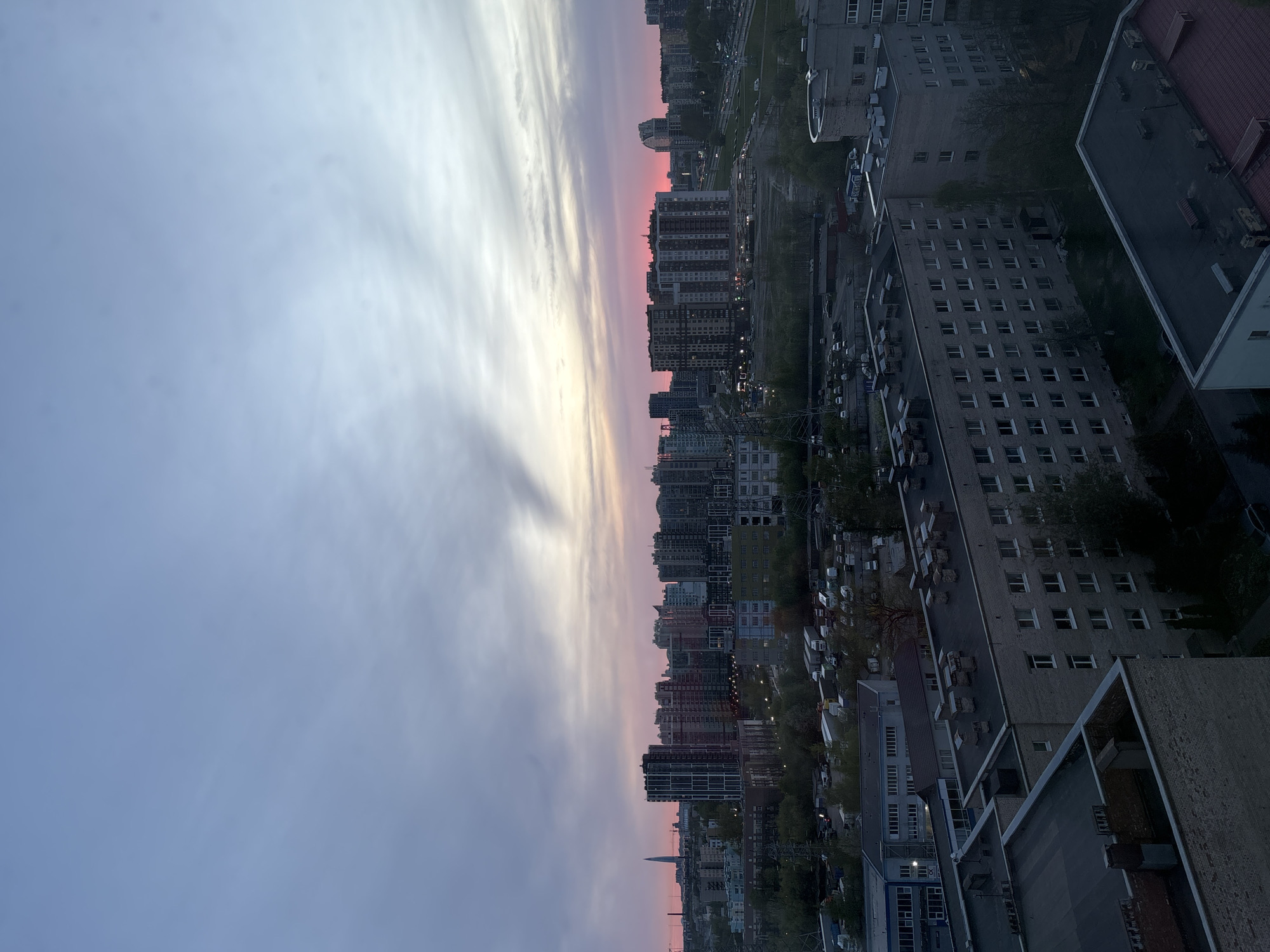 The image depicts a cityscape at twilight. Multiple high-rise buildings line the horizon, silhouetted against a sky graded with colors transitioning from gray to pink and yellow. The urban environment includes densely packed structures and a network of streets and parking areas filled with vehicles. The view is taken from an elevated position, offering a comprehensive perspective of the city below. There are no people visible in the image.