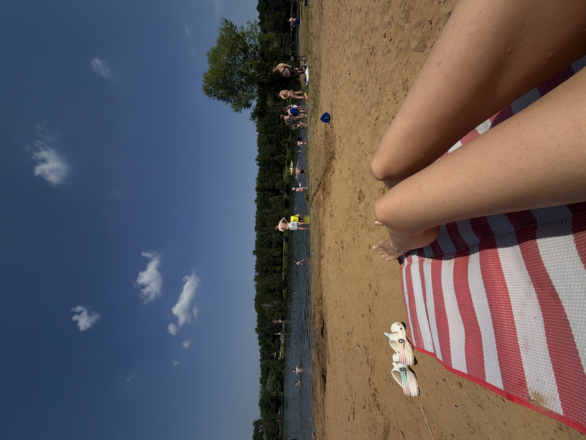 The image depicts a sunny day at a beach, likely in Malakhovka, Russia, with a clear blue sky and scattered white clouds. In the foreground, a person's legs are visible, lying on a red and white striped mat on the sand. Next to the mat are two pairs of white slip-on shoes, one with a rainbow-colored strap and the other decorated with a unicorn horn.
The background features a lake where several people are swimming, wading, and standing. A dense forest lines the far bank of the lake. On the beach, a few individuals are scattered around, with one person standing near the water's edge wearing yellow shorts and a light-colored shirt. A blue bucket is visible in the sand further back from the water. The overall scene conveys a relaxed, summer atmosphere.
