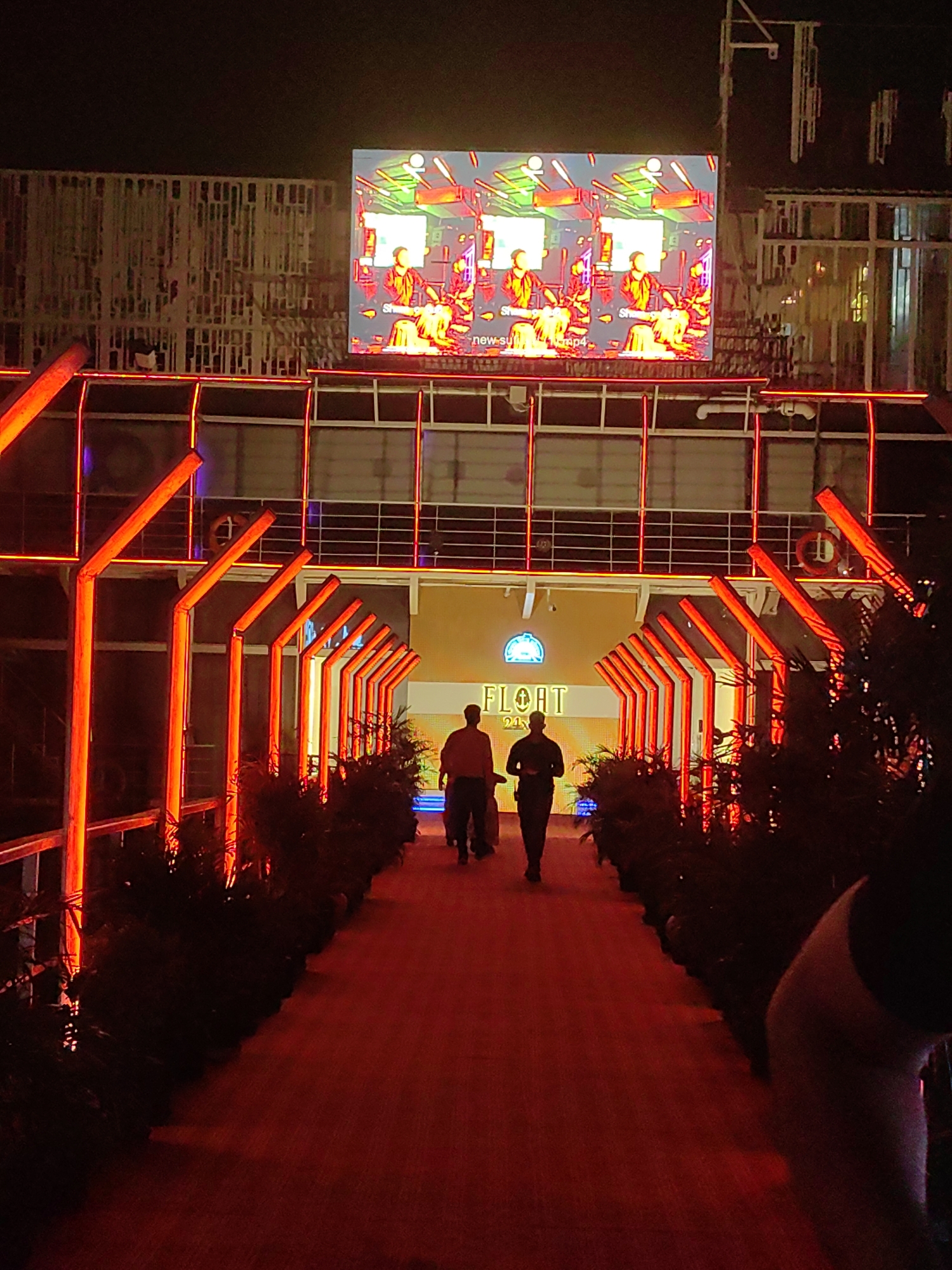 The image depicts a nighttime scene in Gorakhpur, India, featuring a vibrant, illuminated pathway leading towards a building with a large screen displaying a colorful, abstract image. The pathway is bordered by red lights and dark foliage, creating a dramatic and festive atmosphere. Two men are walking away from the camera on the pathway. The building has a yellow facade with the word "FLOAT" and numbers below it in illuminated text. The screen above displays a repetitive graphic with human figures and abstract shapes in bright colors. The overall impression is one of a lively entertainment venue.