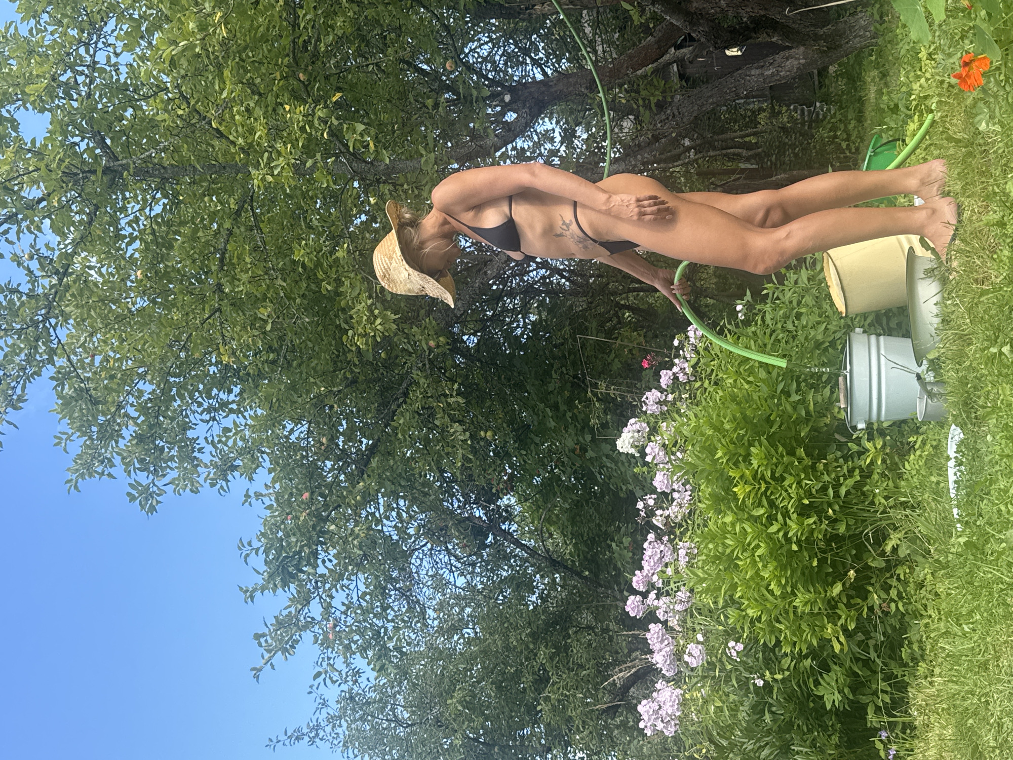 A woman wearing a bikini and a straw hat is standing in a garden. She is holding a green hose. There are trees and flowers in the background. The image appears to have been taken on a sunny day. The woman is partially obscured by foliage, but her legs and torso are visible. There are two buckets and a watering can in the foreground. An orange flower is visible in the upper right corner. The setting suggests a warm day in a rural or suburban area.