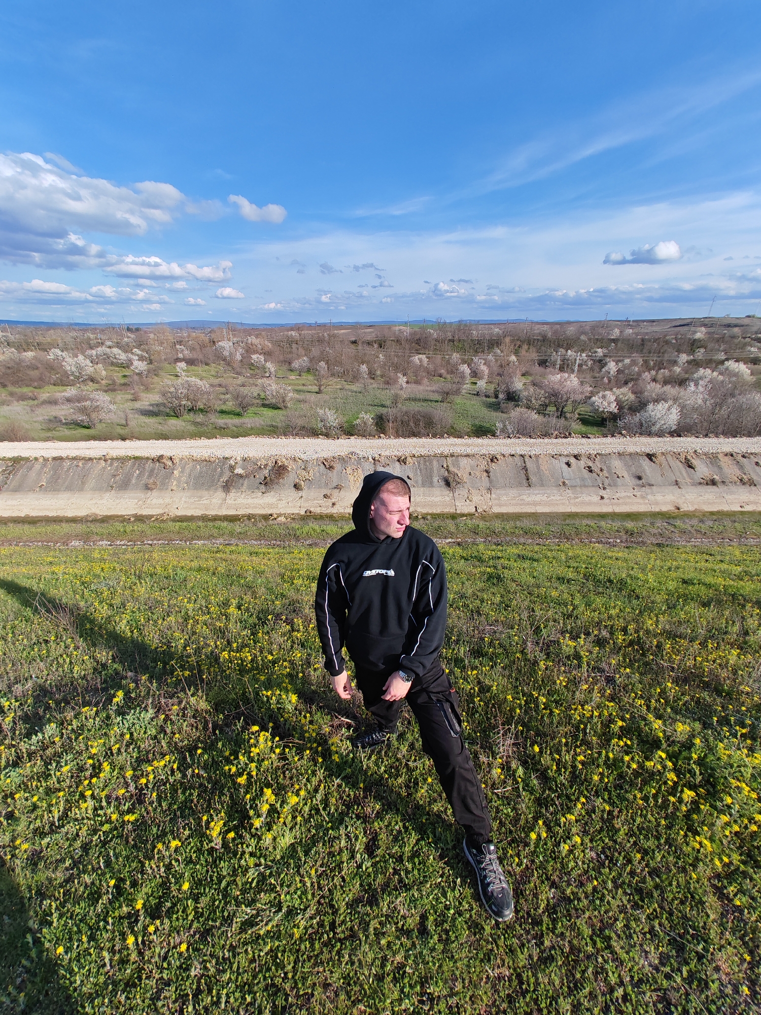 A medium shot shows a man wearing a black hooded sweatshirt and black trousers standing on a grassy hill dotted with yellow flowers. The man is leaning slightly forward, his head turned to the right. Behind him, there's a concrete retaining wall. Beyond the wall, the landscape extends with sparse trees and vegetation under a blue sky with scattered clouds. The photo was taken in Shumen, Bulgaria.
