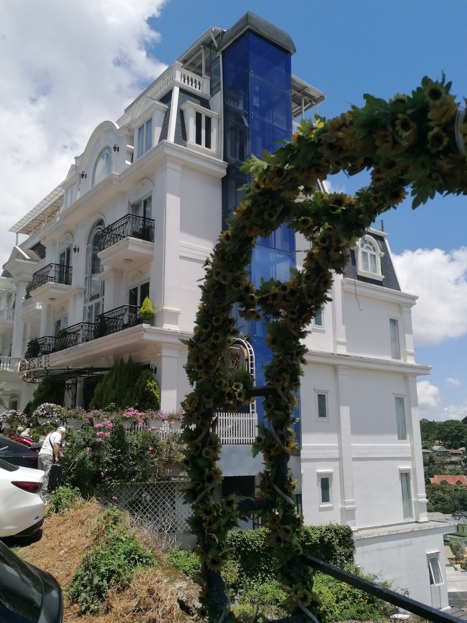 A multi-story white building with a blue glass elevator shaft stands against a bright blue sky with scattered clouds. The building features several balconies with black wrought-iron railings and decorative white trim. A garland of sunflowers and greenery frames the right side of the image. In the foreground, a man in a white shirt and hat is partially visible, carrying a black bag and standing near a white car and some greenery. The building is situated on a hillside, with other structures and trees visible in the background. The word "ENSEE" is visible above the entrance on the first floor. The overall scene suggests a hotel or residential building located in the city of Da Lat, Vietnam, known for its French colonial architecture and hilly terrain.