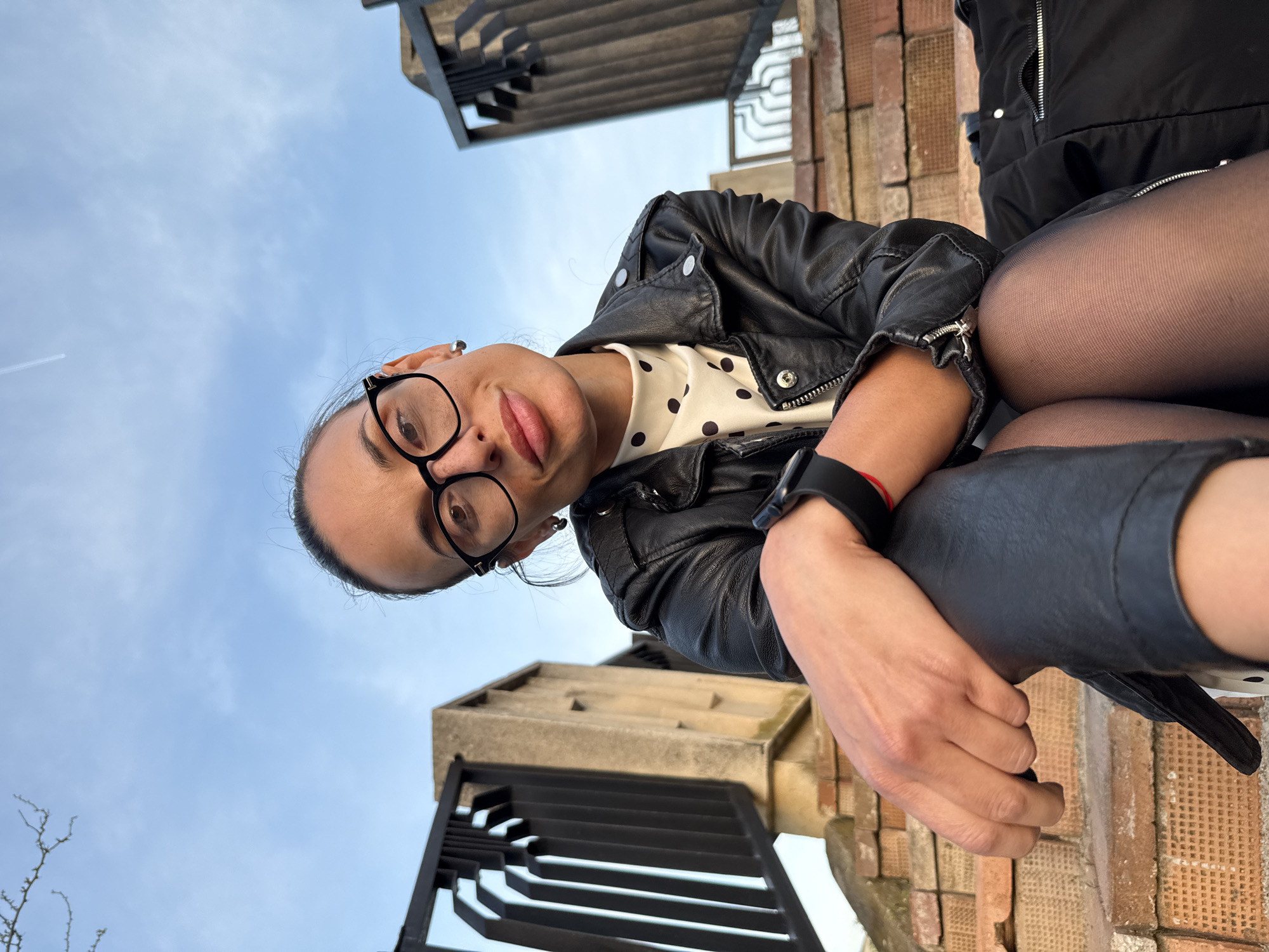 A low-angle, eye-level portrait captures a woman looking directly into the camera with a slightly pouting expression. She has dark hair tied back and wears black-rimmed glasses, which frame her eyes. Her attire consists of a black leather jacket over a white top adorned with black polka dots. On her left wrist, she wears a black smartwatch or fitness tracker and a red bracelet. Her legs, covered in dark tights or leggings, are visible in the lower right section of the frame, suggesting she is seated or leaning back.
The setting appears to be an outdoor urban or architectural environment in Pleven, Bulgaria. The background features a bright blue sky with a distinct airplane contrail visible in the upper left. Architectural elements, including structures made of bricks and concrete, as well as black metal railings with geometric patterns, frame the shot from the top and sides. These elements suggest a public space, possibly a monument or building. The lighting indicates it is daytime, with clear weather. There is no visible text in the image.