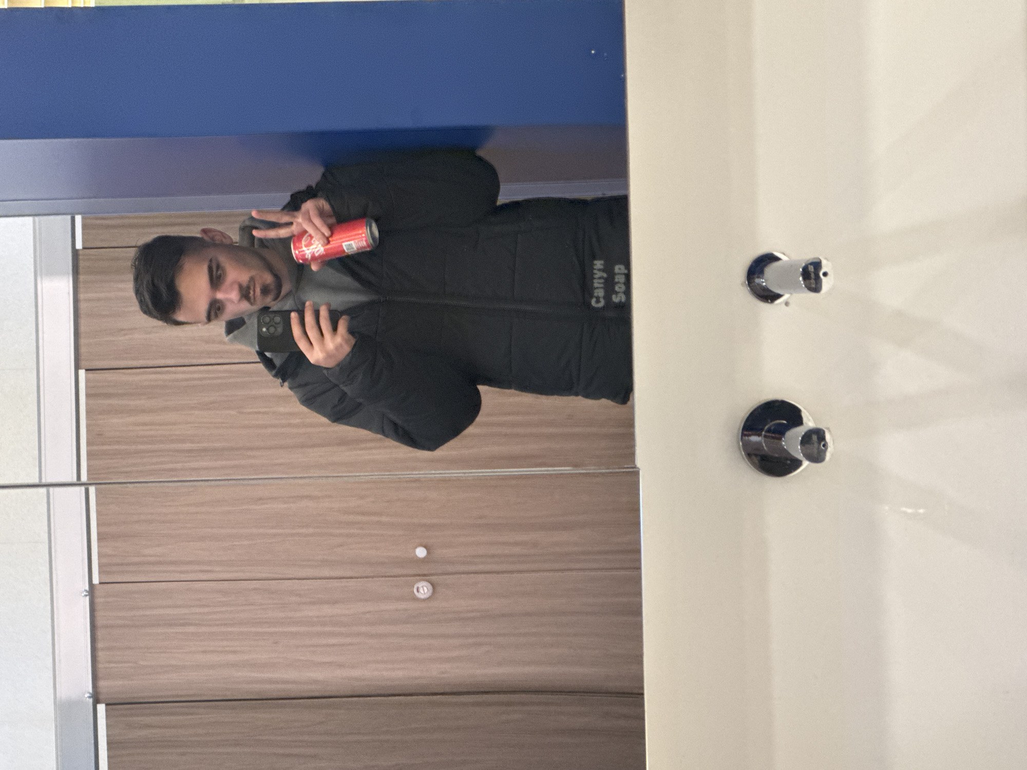 The image is a reflection in a mirror, likely taken with a smartphone. A person is visible in the reflection, holding a beverage can and making a peace sign. The person has dark hair and a beard and is wearing a dark-colored jacket with the text "Сапун Soap" on it. The background in the reflection shows a wall with wooden panels. In the foreground, on the right side of the image, there are two chrome fixtures mounted on a white surface, potentially part of a bathroom or restroom fixture. Based on the text "Сапун" (Soap), the location could be in Bulgaria, and given that Sofia is the capital of Bulgaria, it's possible the image was taken in Sofia.
