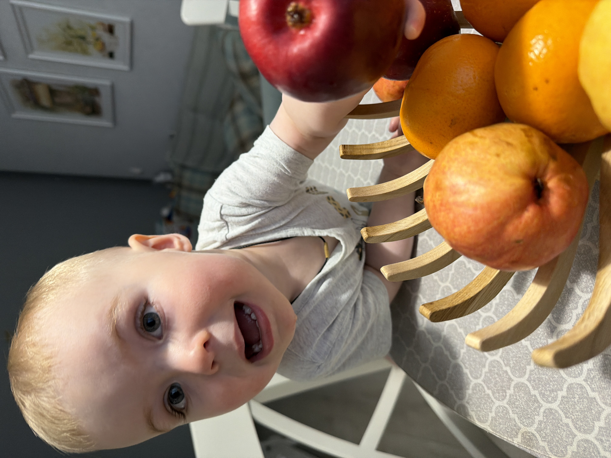 A close-up, upside-down view shows a blond-haired child with blue eyes, possibly between 1 and 3 years old, looking up at the camera with their mouth open. The child is wearing a light grey shirt. Above the child is a fruit basket filled with red apples and oranges. The basket's wooden frame is visible. The child appears to be holding the basket. The background reveals a patterned tablecloth on a white table, and in the blurred distance are framed pictures on a grey wall. The location is unlikely to be Votkinsk, Russia due to the unlikelihood of the photo including geographical signifiers.