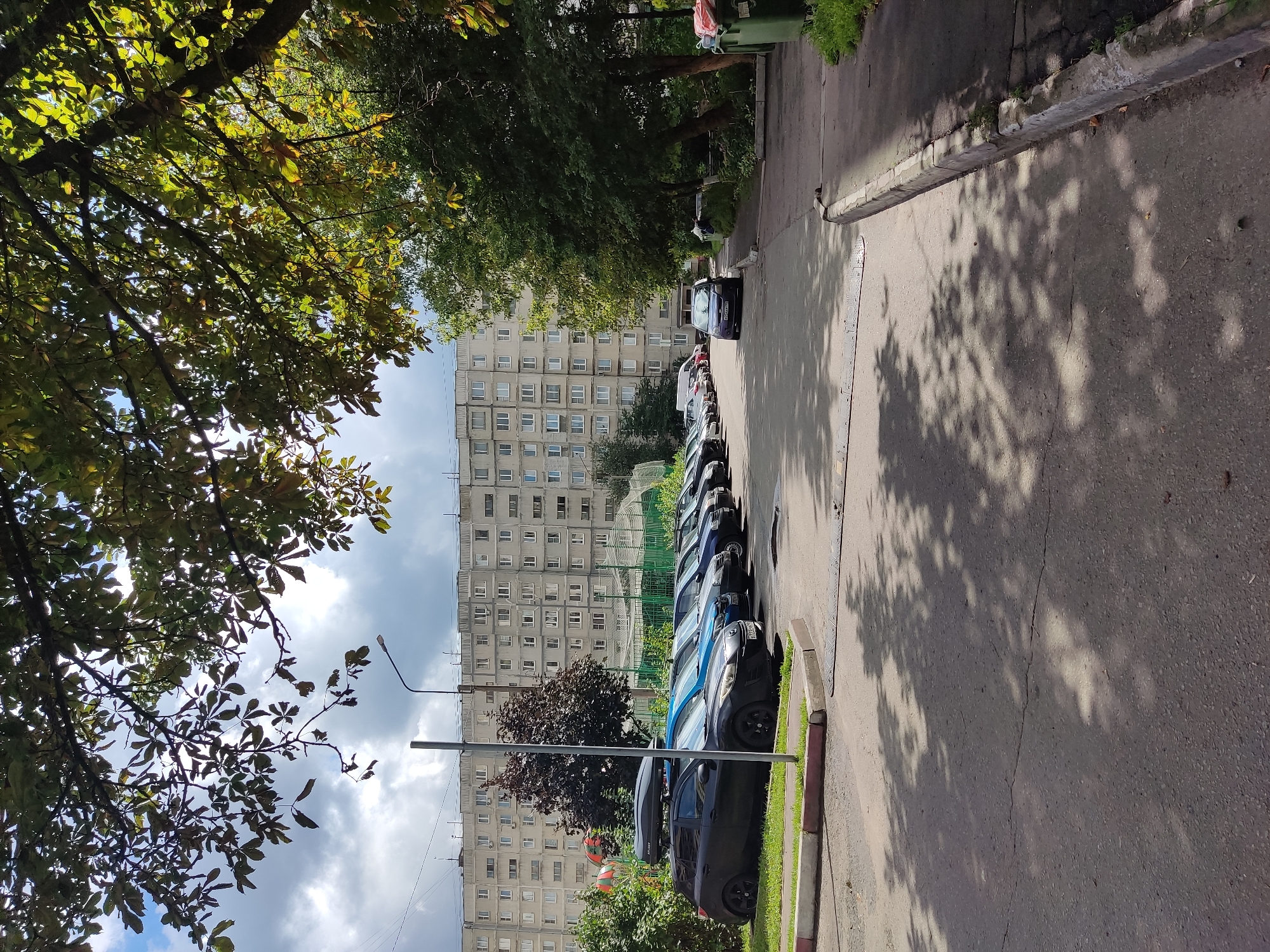 The image shows a parking lot in front of a multi-story residential building. Several cars are parked in a row. A dark SUV with a roof box is parked on the left, followed by a blue hatchback, and then a series of darker sedans and hatchbacks. A purple sedan is parked further down the lot, closer to the building. Trees with green and yellowing leaves overhang the parking area, casting dappled shadows on the asphalt. The building appears to be an older, concrete structure with numerous windows. A street light stands near the left edge of the frame. In the background, there is a chain-link fence and some greenery. The sky is partly cloudy with patches of blue. Two green garbage bins are visible on the right side of the image. The scene is consistent with an urban environment in Noginsk, Russia.