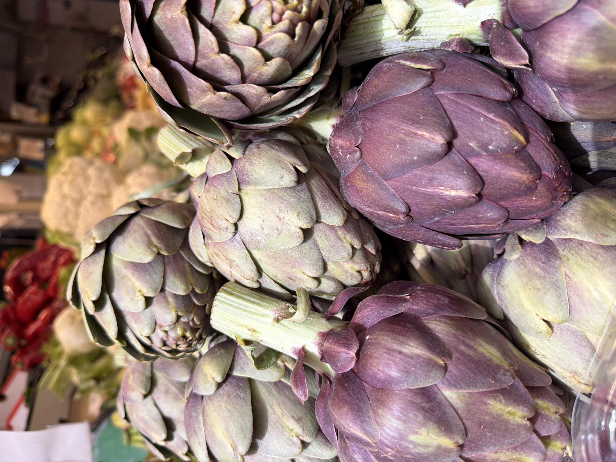 A close-up, high-angle shot displays a cluster of artichokes. The artichokes vary in color, ranging from green to purple hues. In the background, there are other vegetables, including what appears to be cauliflower. The setting is likely a market or grocery store in Tel-Aviv, Israel. The focus of the image is on the texture and colors of the artichokes.