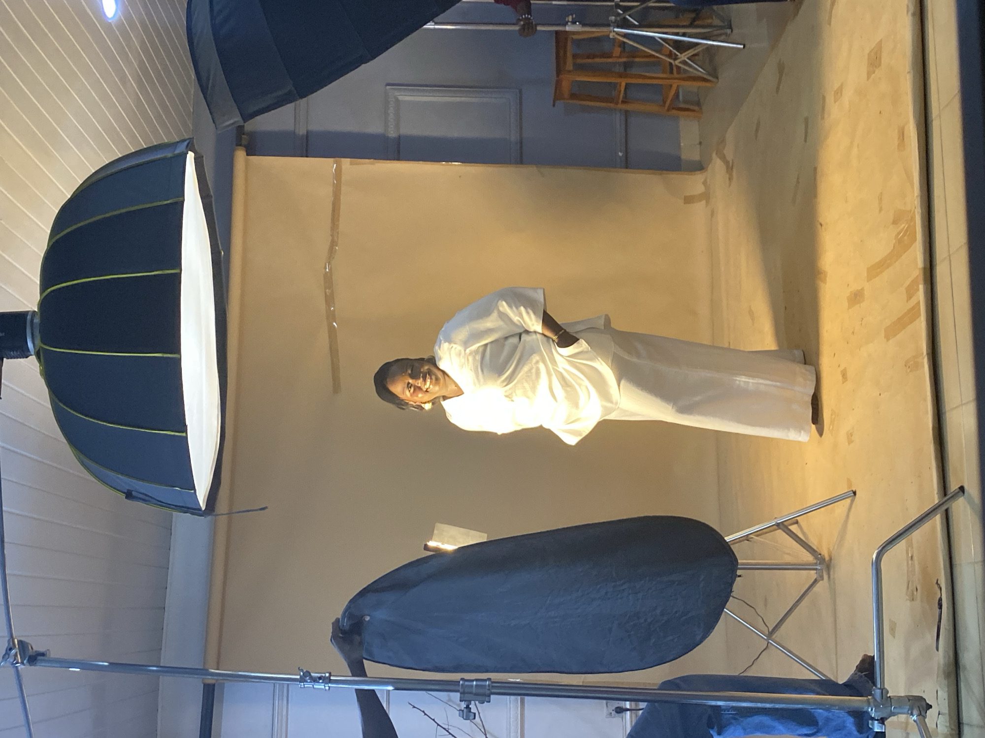 A person in a white outfit stands in a room with a beige backdrop. The person is facing left and looking at the camera with a slight smile. The person is wearing a headscarf and appears to be in a studio setting, as indicated by the presence of professional lighting equipment. A large softbox light is positioned to the left of the person, and a reflector is held by a person whose hands are visible at the bottom of the frame. The backdrop is made of fabric and is taped to the wall. A wooden chair is visible in the background, as well as some studio equipment. The room appears to be lit by artificial lights, and there are no windows visible, so the time of day and weather cannot be determined. The overall mood of the image is calm and professional, suggesting a photoshoot is in progress. The location context is Bukuru, Nigeria, but there are no specific visual cues in the image to confirm this. There is no visible text in the image.