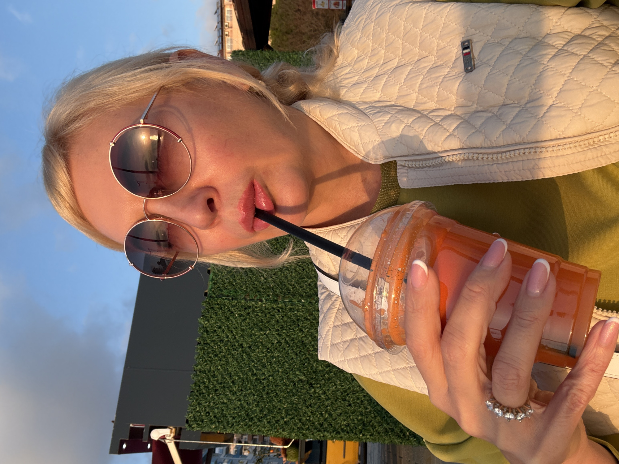 The image is an upward-angled, close-up portrait of a person, likely female, with blonde hair, captured outdoors during what appears to be the golden hour or sunset. The warm, directional light illuminates the subject's hair and clothing.
The subject is wearing large, round sunglasses with reflective lenses that show the bright sun, a blue sky, and hints of clouds. Their lips, adorned with pink lipstick, are pursed around a black straw, from which they are drinking an orange-colored beverage from a clear plastic cup.
The person is dressed in a light-colored, possibly cream or beige, quilted vest worn over a green top. A small, rectangular emblem featuring red, white, and blue stripes is visible on the quilted fabric of the vest. The subject's right hand, holding the drink, has nails painted in a French manicure style, and a ring with several clear, sparkling stones is worn on the middle finger.
The background includes an artificial green hedge and blurred structures, suggesting an outdoor setting such as a patio, rooftop, or cafe. The clear blue sky is prominent above the hedge and buildings. No discernible text is visible other than the emblem on the jacket, which does not contain legible letters.