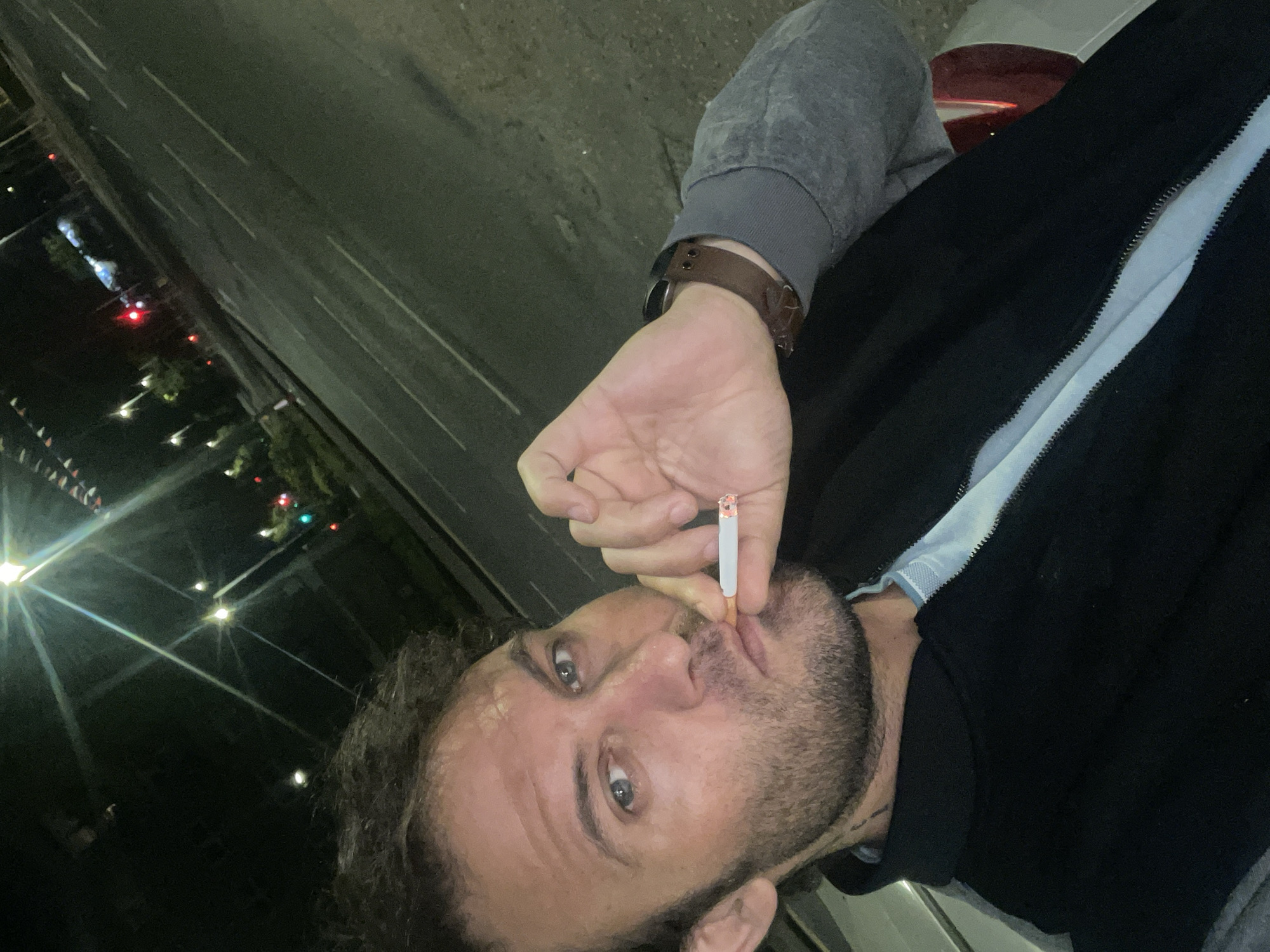 A man is holding a lit cigarette to his mouth, mid-puff, in a close-up selfie. He has short, dark, messy hair, blue eyes, and stubble. He is wearing a black zip-up jacket over a light blue collared shirt. A brown watch is visible on his left wrist. The background is dark and appears to be a street at night, with streetlights and traffic lights visible. The lighting suggests this is an urban environment. The man is positioned in the foreground, with the street and lights in the background, out of focus. The image is taken from a slightly elevated angle, looking down towards the man. The presence of streetlights and what appear to be traffic signals suggests a road in a town or city. Given the prompt's mention of Armagh, United Kingdom, this scene likely depicts a street within that city.