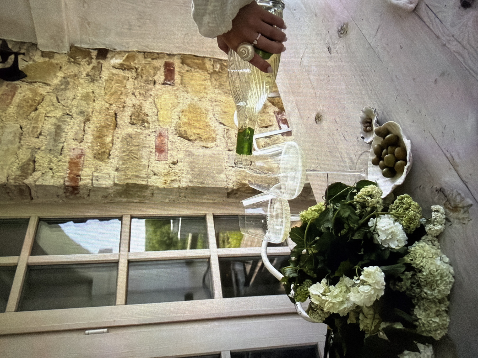 The image captures a moment of beverage pouring in what appears to be a rustic yet elegant setting, located in Ros Na Rí, Ireland. The scene is presented from a slightly elevated, overhead perspective, with the original image rotated ninety degrees counter-clockwise.
In the center of the frame, a person's hand is visible, clad in a white or off-white long-sleeved garment, holding a clear glass bottle. A light-colored, possibly beaded, ring adorns the middle finger of the hand. The bottle, containing a light-colored liquid (likely white wine), has a green foil or label towards its base, and is tilted to pour into one of two empty clear wine glasses below. These glasses are adorned with translucent white ribbons tied around their stems.
To the right, on a light-colored, possibly painted wooden surface, sits a small, white, organic-shaped bowl filled with green olives. Another smaller, similar bowl with a few olives is next to it. In the foreground, a substantial arrangement of white and light green hydrangeas or similar cluster flowers, mixed with dark green foliage, is displayed in a partially visible white vase or container.
The background features a rustic stone wall composed of irregular yellowish stones and scattered reddish bricks. To the lower left, a large multi-paned window or glass door with light-colored wooden frames reflects greenery from outside, indicating daylight conditions. A dark, metallic, wall-mounted object is partially visible on the stone wall to the far left.
The overall atmosphere suggested by the elegant flowers, wine, and olives, combined with the rustic architecture, is one of a relaxed social gathering or preparation for such an event. No visible text is present in the image.