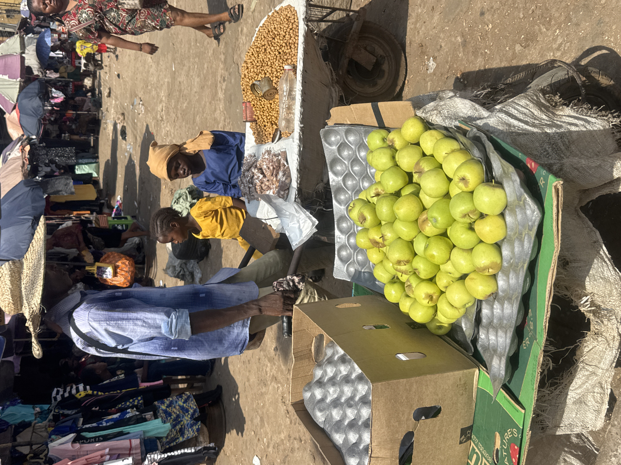 This image captures a lively outdoor market scene, likely in Onitsha, Nigeria, under bright daylight. The ground is unpaved and appears dusty.
In the foreground, a man wearing a light blue patterned long-sleeve shirt, dark pants, and a straw hat draped over his back stands, facing towards other market activities. His dark hand is visible.
Central to the image are two individuals, possibly vendors, positioned behind a makeshift stall. One wears a dark blue top and a light brown head covering, while a younger person in a yellow shirt sits beside them. Their stall prominently displays a large mound of light brown, round items, likely groundnuts or chickpeas, with a clear plastic bottle and an empty tin can placed among them. Below this, there's a smaller pile of darker, reddish-brown produce.
Adjacent to this stall, a cardboard box is filled to the brim with numerous bright green apples, which are arranged in grey, egg-tray-like protective inserts. Part of the box is visible, showing text that includes "NATURE'S" and "SOUTH A" (likely referring to South Africa). Another open cardboard box with similar grey inserts is nearby, alongside a large, loosely woven burlap sack.
In the background, a bustling market environment is visible with numerous stalls, clothes hanging on racks, and umbrellas providing shade. A woman in a colorful patterned dress walks through the market. The strong sunlight casts distinct shadows across the ground and objects, indicating a clear, sunny day. The overall scene depicts typical daily commerce and activity within an open-air market.