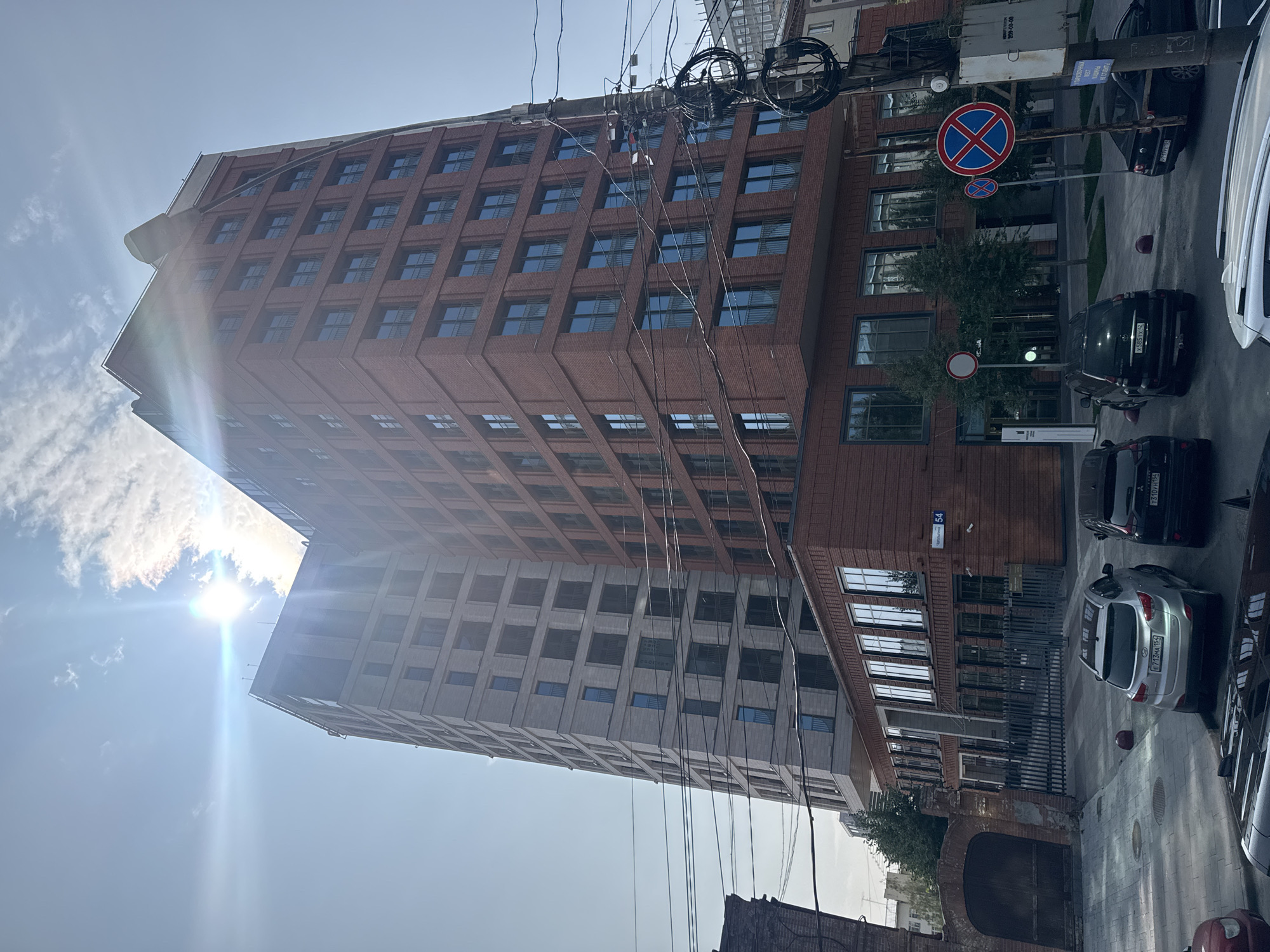 The image shows a modern brick building in Novosibirsk, Russia, with a lighter colored building adjacent to it. The sun is shining brightly, creating lens flare. Several cars are parked in front of the buildings, and there are power lines and traffic signs in the foreground. The street number "54" is visible on the brick building.