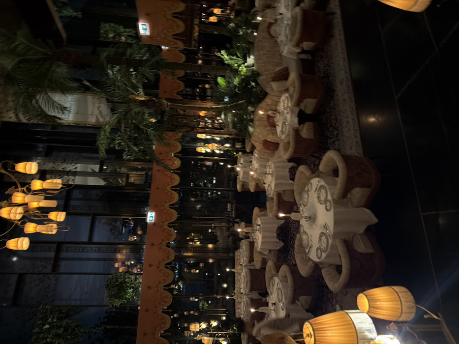 The image displays the luxurious interior of La Cantine du Faubourg, located within Jumeirah Emirates Towers in Dubai, United Arab Emirates. The scene is set at night, indicated by the dark exterior visible through expansive, dark-framed windows that line a significant portion of the far wall.
A large, ornate brass chandelier featuring multiple amber, fringed lampshades hangs centrally from the ceiling, illuminating the space with a warm glow. Below the chandelier, a decorative orange-gold valance or awning with intricate trim extends horizontally across the window section. Green "EXIT" signs are visible above this valance.
The dining area is filled with numerous circular tables, each draped with a white tablecloth and prepared with white plates featuring dark concentric patterns, glassware, and cutlery. A lit candle in a holder is present on each table. Seating comprises plush, upholstered armchairs in a light cream or beige hue, often accented with a darker, reddish-brown outer shell, and curved banquettes or sofas in similar light upholstery. The floor is covered by a patterned rug or carpet, with a darker, possibly tiled, floor visible in the immediate foreground.
Several large, potted palm trees and other green foliage are strategically placed throughout the restaurant, contributing to a lush, garden-like atmosphere. The primary lighting sources are the ceiling chandeliers, table lamps, and individual candles, creating a warm, ambient illumination. One blurred individual, appearing to be walking, is partially visible in the mid-left portion of the frame. The restaurant appears largely unoccupied by diners at the time the image was captured.