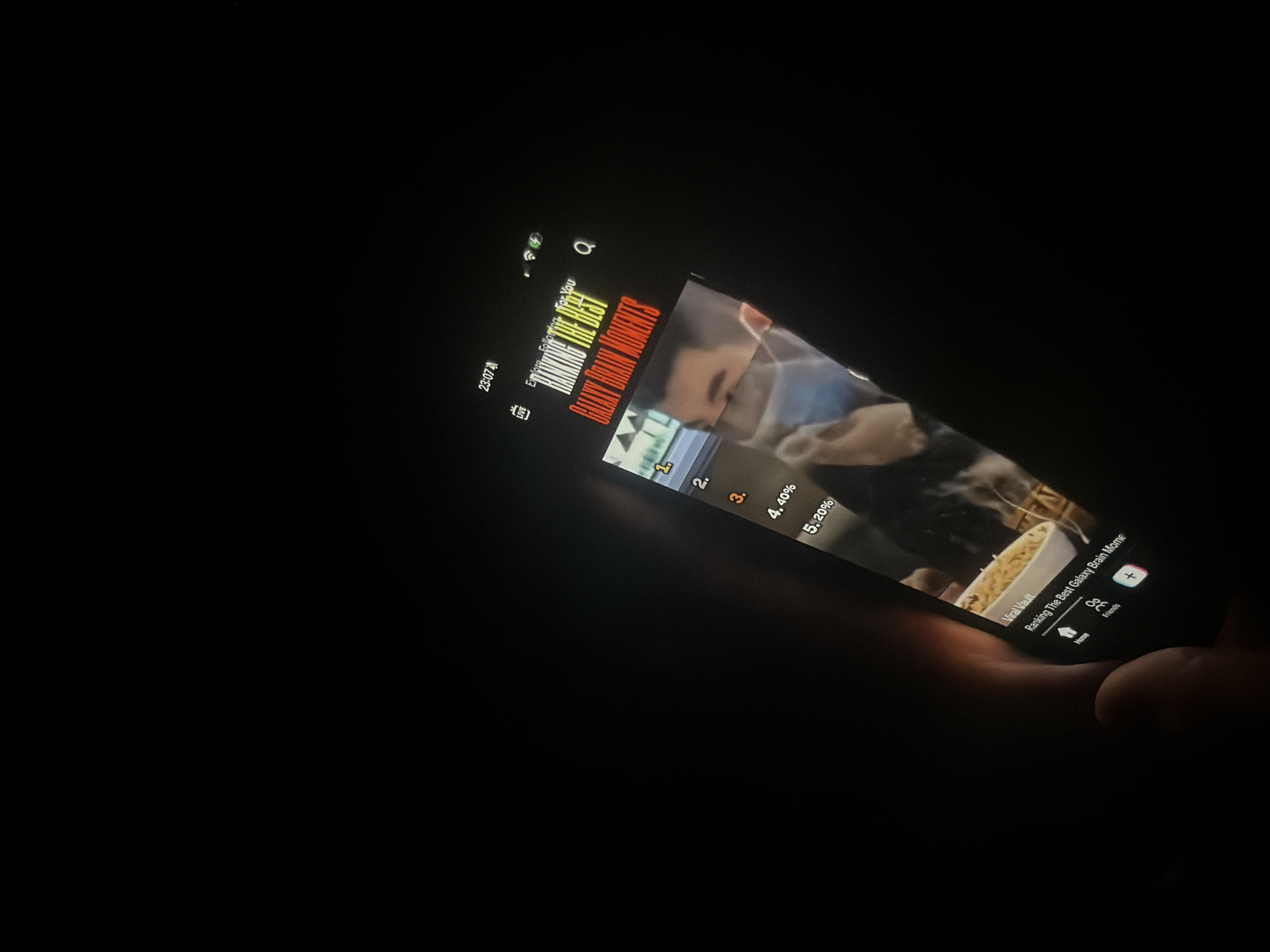 The image captures a smartphone being held in a dark environment, likely a room at night. The glowing screen is the primary source of light, illuminating parts of a hand holding the device. The phone's display shows content from what appears to be a video-sharing or social media application.
The visible content on the screen features a video or image with a person, possibly a man, and a white bowl containing what looks like noodles. Overlaid text indicates a ranking or list, with the main title reading ""RANKING THE BEST" GALAXY BRAIN MOMENTS". Smaller text below the video thumbnail reads "Viral Vault" and "Ranking The Best Galaxy Brain Mome". A list of items is visible, partially obscured, showing "1.", "2.", "3.", "4. 40%", and "5. 20%", suggesting a percentage-based ranking.
On the top left of the screen, the time "23:07" is displayed, indicating late evening or night. Other user interface elements visible include "LIVE", "Explore", "Following", "For You", a search icon (Q), and navigation options at the bottom such as "Home", "Friends", and a plus icon. The overall scene suggests a person consuming digital media in a dimly lit setting.