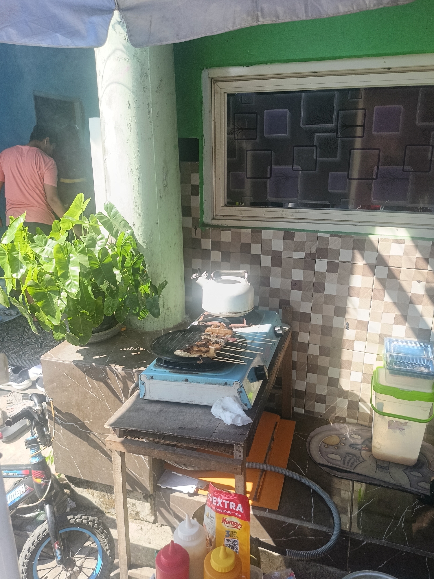 An exterior shot captures a scene in Gedangan, Indonesia, featuring a man in a pink t-shirt cooking outdoors. He is positioned behind a large, leafy green plant and is partially obscured by smoke. In the foreground, a small blue and white gas stove is set up on a wooden table, with skewers of food cooking on a grill. A white kettle sits on the stove. Nearby, a bicycle is parked, and several containers, including a large white bucket with a green lid, are visible. The wall behind the cooking area is tiled with a checkered pattern of brown and beige squares. A window with a patterned film on the glass looks out onto the scene. The overall setting appears to be a residential outdoor space.