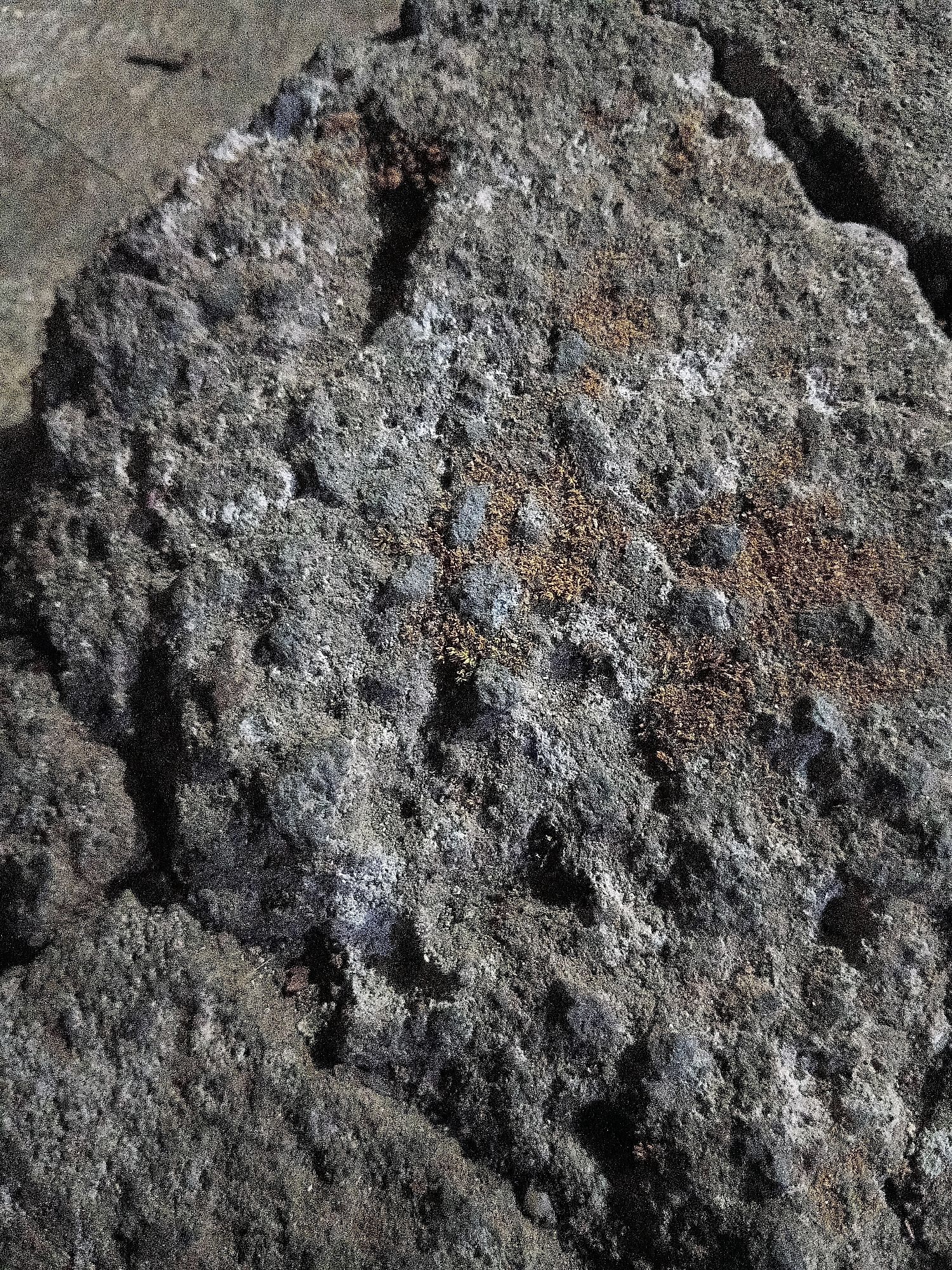 The image shows a close-up, high-angle view of a rough, textured rock formation. The rock is predominantly dark gray with patches of lighter gray and some areas of reddish-brown mineral deposits or moss. The surface is uneven, with visible pores, crevices, and protruding fragments, giving it a porous and somewhat weathered appearance. The lighting creates strong shadows, highlighting the rock's irregular topography. The background is out of focus and appears to be a gritty, earthy surface, possibly more rock or soil. There are no people, animals, or man-made objects clearly visible in the frame. The scene conveys a natural, geological texture. The location within the city of Cikal, Indonesia is not discernible from the image itself.