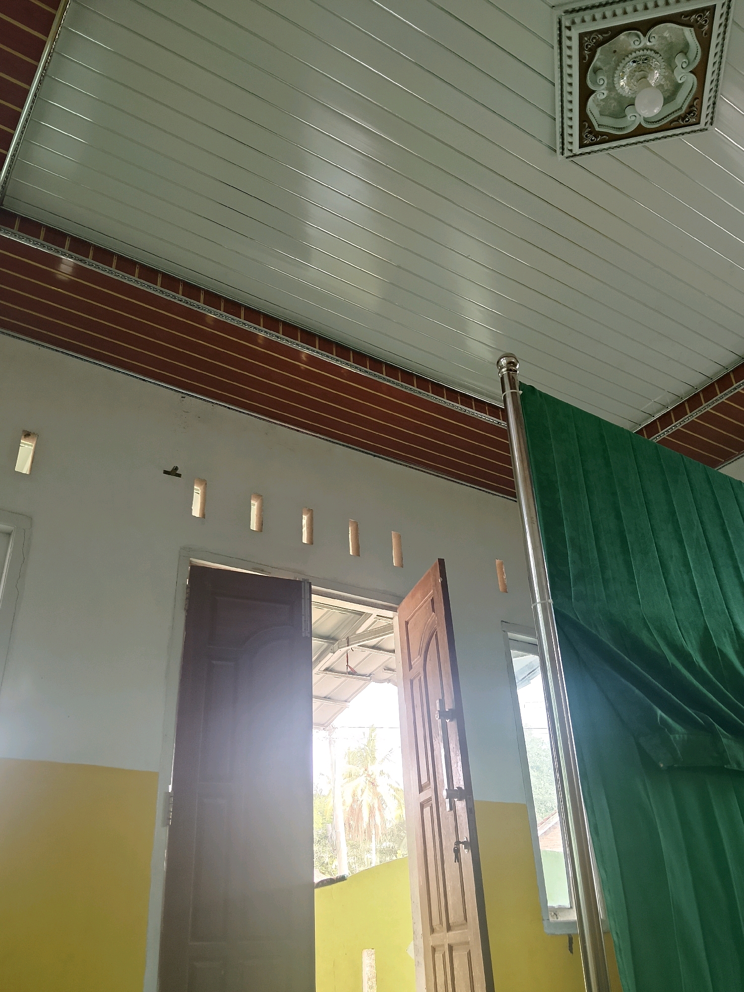 This image shows an interior view of a room, likely in a residential building in Negeri Agung, Indonesia. The room has a white ceiling with a decorative light fixture and horizontal brown trim. The walls are painted white and a portion of the lower wall is painted yellow. There are several rectangular openings in the wall, possibly for ventilation. Two wooden doors are visible, one open and one partially open. A tall, silver pole stands near the open door, draped with a green curtain. Sunlight streams through the open doorways and windows, creating lens flare. In the background, through the open doors and windows, palm trees and other foliage are visible, suggesting a tropical outdoor environment. There are no people in the image.