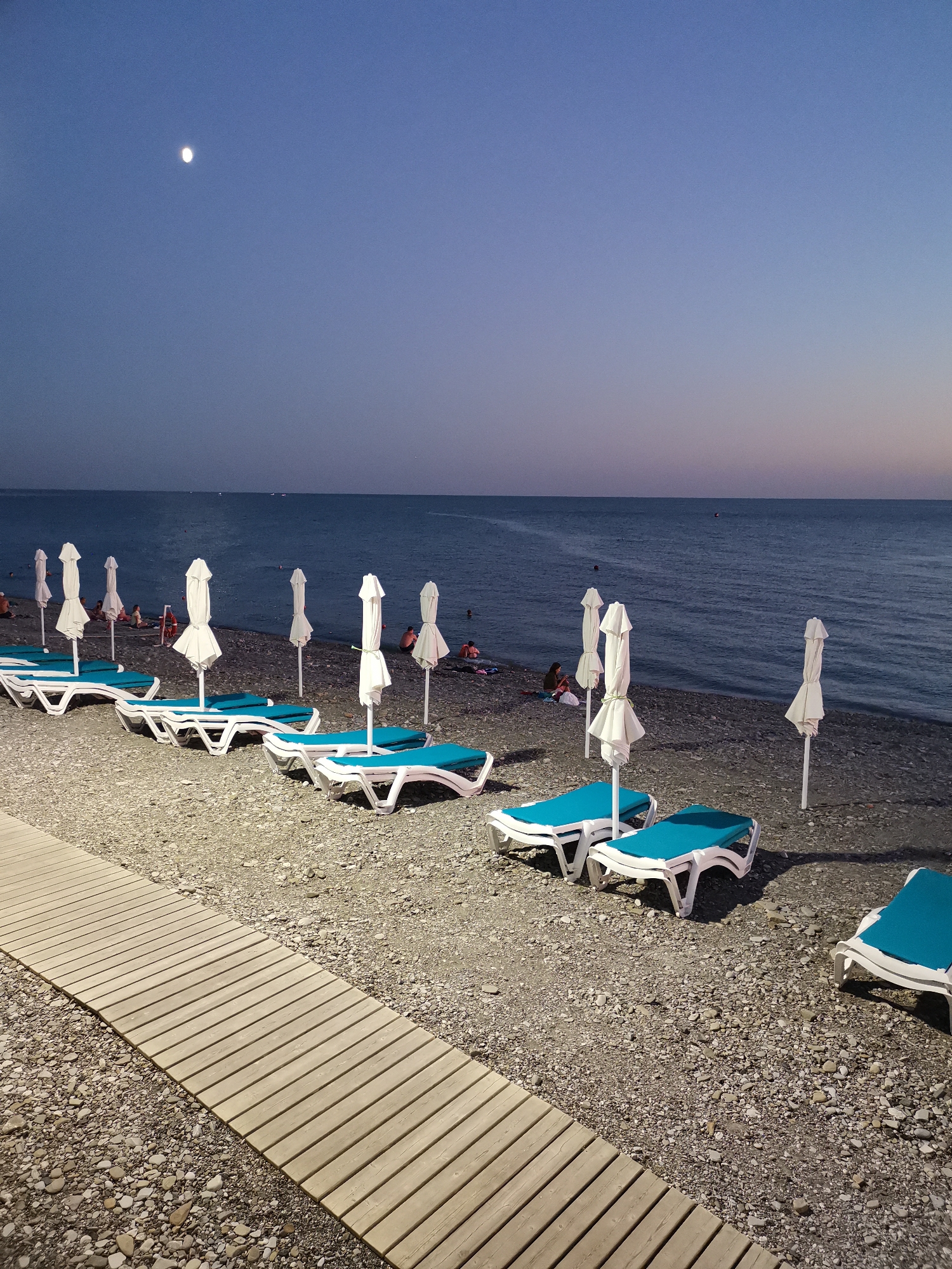 The image depicts a beach scene in Khostinskiy, Russia, at dusk or night. A wooden boardwalk runs diagonally from the lower left corner towards the center of the image. Beyond the boardwalk, a pebble beach is lined with numerous white beach umbrellas and blue-cushioned lounge chairs, most of which are unoccupied. In the background, the dark blue ocean stretches to the horizon, with a faint glow of twilight in the sky. A bright full moon is visible in the upper left quadrant of the sky. A few people are scattered on the beach and in the water in the distance, appearing as small figures. The overall atmosphere is calm and serene.