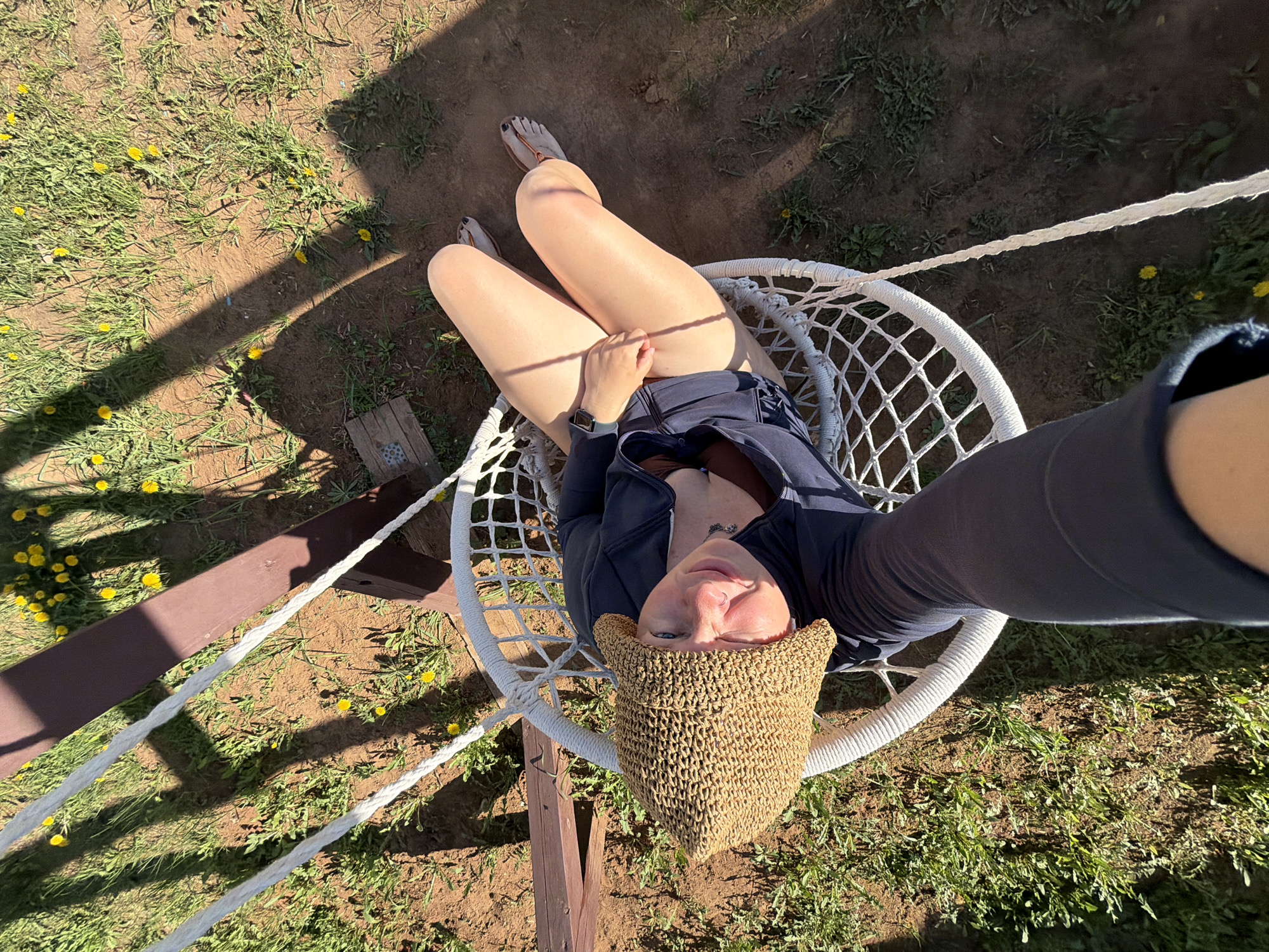 A high-angle, full-body shot captures a fair-skinned person in a round, white, macrame-style swing, likely outdoors. The individual wears a dark zip-up jacket, shorts, and a straw sun hat. Their legs are crossed, with a digital wristwatch visible on the wrist of the hand resting on their thigh. The person appears to be holding a smartphone or similar device to take the photo. The ground around the swing is a mix of dry dirt and grass with scattered yellow dandelions. Support beams for the swing are visible. Based on image geolocation data, the scene is located in Chaikovsky, Russia.