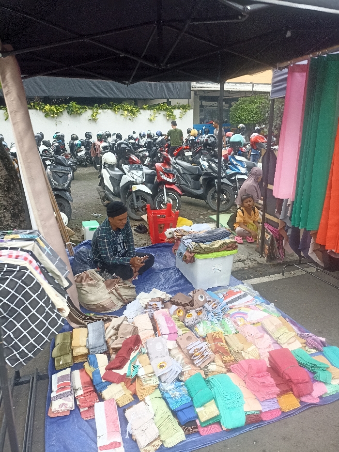 The scene is a street market in Purwokerto, Indonesia, located at latitude -7.418 and longitude 109.253. A man wearing a checkered shirt and a black cap is sitting on the ground under a canopy, looking down at his phone. He is surrounded by stacks of fabric arranged in a pattern on a blue tarp, while several other people are standing around him, including a young girl with a pink scarf and a woman with a white scarf. The man appears to be selling fabric. In the background, a large number of motorbikes are parked, facing a white wall with an open area behind it. The ground is a gray, paved surface, with a few fallen leaves visible. There are some items hanging on a string of rope on a side stand, and one person appears to be assisting with the organization of the fabric.