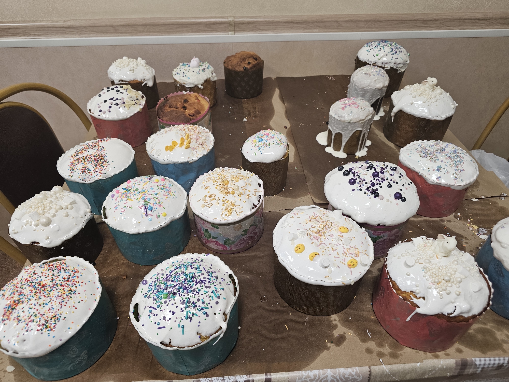 The image shows a collection of decorated Easter cakes (kulich) arranged on a table covered with baking paper and a tablecloth. The cakes vary in size and decoration, with white icing and sprinkles of various colors and shapes. Some cakes also have edible decorations like small yellow faces and white bunny figurines. The cakes are baked in paper molds that come in a variety of colors and patterns. A chair is visible on the left side of the image. The setting appears to be indoors, possibly in a home kitchen or dining room. The location is not evident from the image.