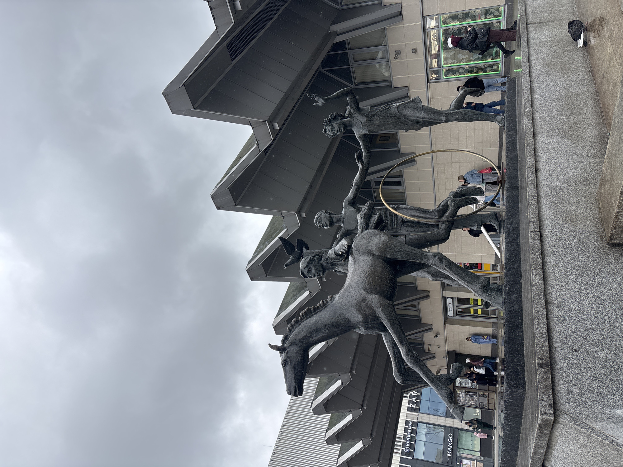 The image depicts a bronze statue in front of a modern building in Saint Petersburg, Russia. The statue features a horse with riders and a hoop. Several people are walking nearby. The sky is overcast. Businesses can be seen in the background.