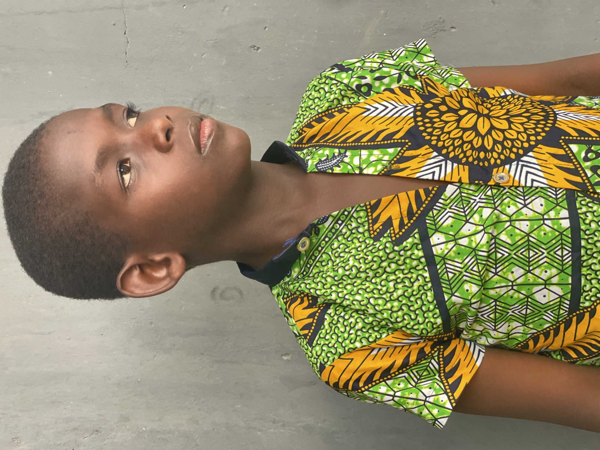 A young boy with dark skin and short, dark hair is depicted in a close-up, chest-up shot. He is looking upwards and slightly to his right with a calm, contemplative expression. His eyes are open, and his lips are lightly colored.
He is wearing a vibrant, short-sleeved button-up shirt featuring a traditional African print, often referred to as Ankara fabric. The pattern is characterized by a striking combination of bright green, golden yellow, black, and white. Prominent design elements include large, circular yellow motifs resembling sunbursts or flowers on a black background, intricate geometric patterns in green and white, and swirling or dotted patterns. The shirt has a dark-colored collar and small, gold-toned buttons down the front.
The background is a plain, light grey, slightly textured surface, possibly a concrete wall or floor. There are minor cracks and imperfections visible on this surface, indicating an unadorned setting. The lighting appears soft and diffused, suggesting an indoor or shaded outdoor environment, without harsh shadows. The boy's attire and the provided context indicate the location is Badagry, Nigeria. There is no visible text in the image.