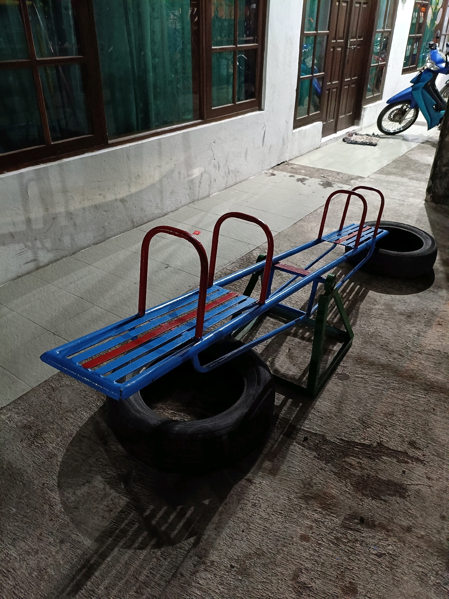 The image depicts a blue and red seesaw situated outdoors on a paved surface. The seesaw is supported by a green metal frame and rests on two large tires, one on either side. The background shows a building with wooden-framed windows and doors, some of which are covered by green curtains. To the right of the building, a blue motorcycle is parked. The scene is illuminated by artificial light, casting shadows from the seesaw onto the ground. The location is identified as Maguwoharjo, Indonesia.