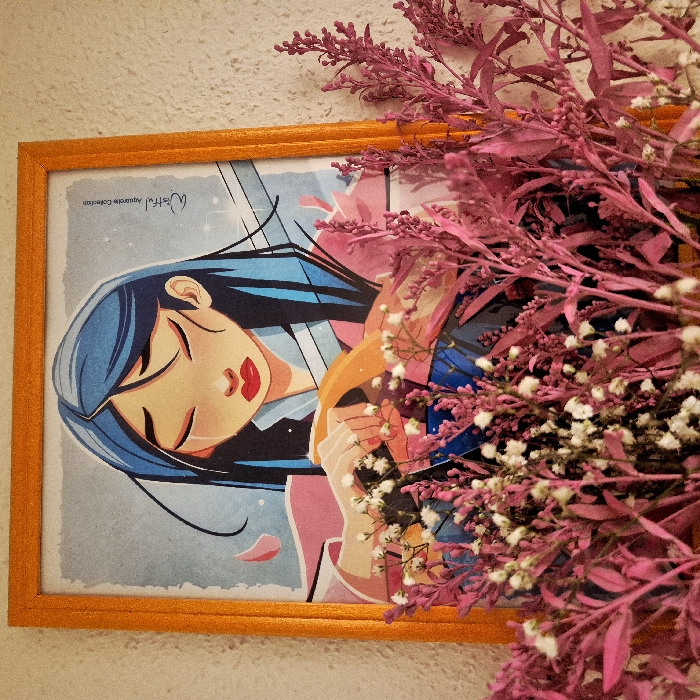 A vertically oriented illustration, contained within a glossy orange frame, is mounted on a light-colored, textured wall in Strasbourg, France. The artwork depicts a female character with light skin, blue-black hair, closed eyes, and prominent red lips, with tear trails visible beneath her eyes. She wears a pink upper garment over a darker underlayer, with her hands resting on a curved yellow object, possibly a sword hilt. The background of the illustration is a speckled light blue, featuring several floating pink petals. The text "Wishful" and "Aquarelle Collection" is visible in the upper right area of the print. The lower portion of the framed artwork is partially obscured by a dense cluster of floral elements. This cluster consists of numerous small white flowers, characteristic of Baby's Breath, intermingled with tall, thin sprigs of magenta-colored dried botanical elements.