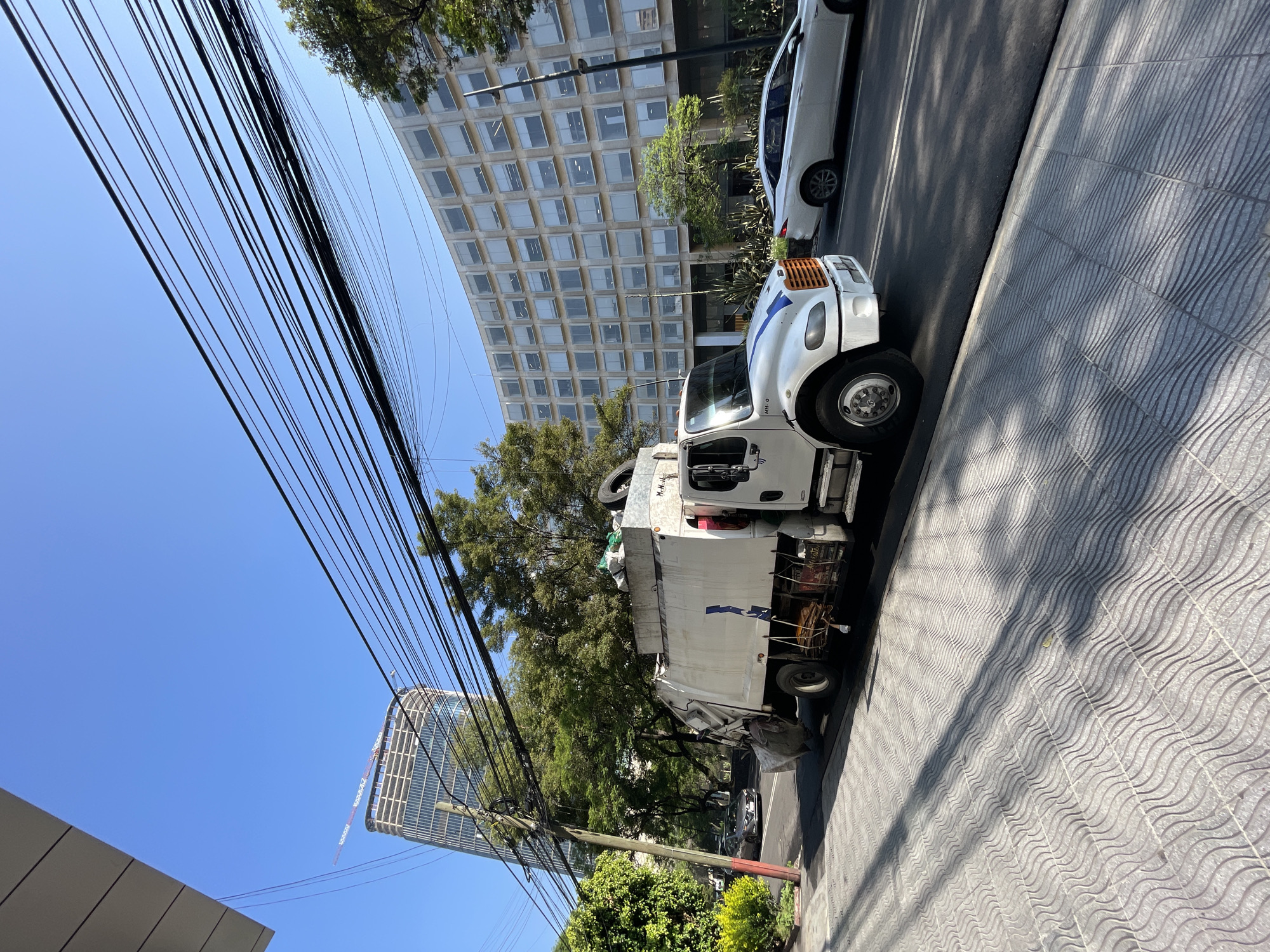 The image depicts a street scene in Mexico City, Mexico, on a sunny day. A white garbage truck is parked on the side of the road, casting a shadow on the patterned sidewalk. A white sedan is parked behind the truck. In the background, tall office buildings with grid-like window patterns are visible. A cluster of power lines is prominent in the foreground, obscuring part of the sky and buildings. The foliage of several trees adds greenery to the urban landscape. The overall impression is an everyday street scene.