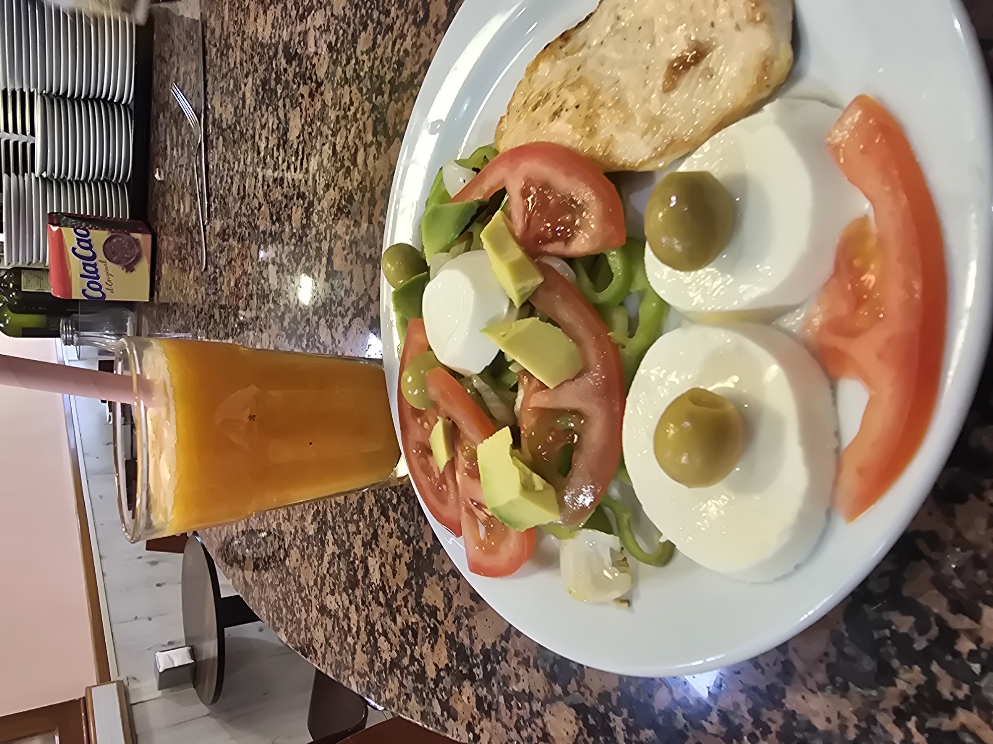 An overhead shot depicts a bright orange beverage in a clear glass, alongside a salad with a piece of grilled chicken and a slice of tomato, arranged in the shape of a smiley face. The food is served on a white plate, resting on a granite tabletop. In the background, a stack of white plates and a small yellow box labeled "Cola Cao" are visible. The scene appears to be in Barcelona, Spain, possibly at a cafe or restaurant. The lighting is bright, casting subtle shadows and highlighting the textures of the food and tabletop.