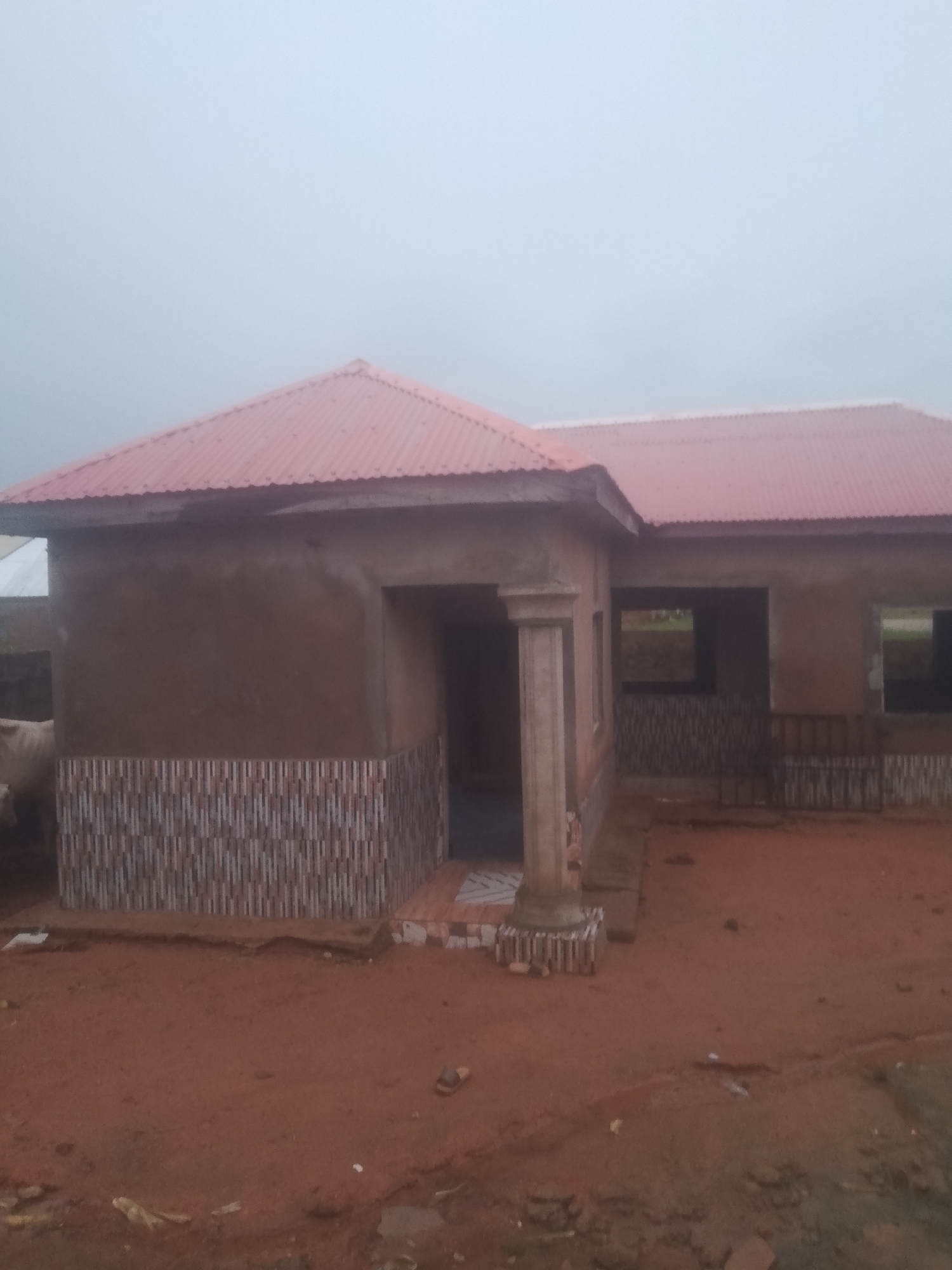 A house under construction in Jauro Yinu, Nigeria, features a red corrugated metal roof, brown plastered walls, and a tiled base. The house has an entrance with a pillar and an open doorway. Windows are visible, some with dark frames. The ground around the house is red earth with scattered debris. A cow is partially visible on the left side of the image. The sky is overcast.