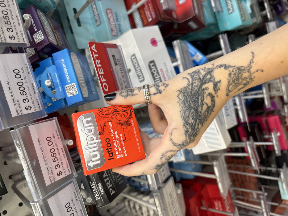 A tattooed hand selects a red "Tulipan Tattoo" condom pack from a shelf filled with various condom brands. The price tags nearby show prices ranging from 3,500 to 7,410. The hand features black nail polish and multiple rings on the ring finger. Condom brands like "Tulipan" and "Prime" are visible, with the "Tulipan" brand appearing in multiple variations and pack sizes. Some packs have "Oferta" stickers, indicating a sale. The shelf displays a variety of colors and product descriptions, suggesting a range of options.