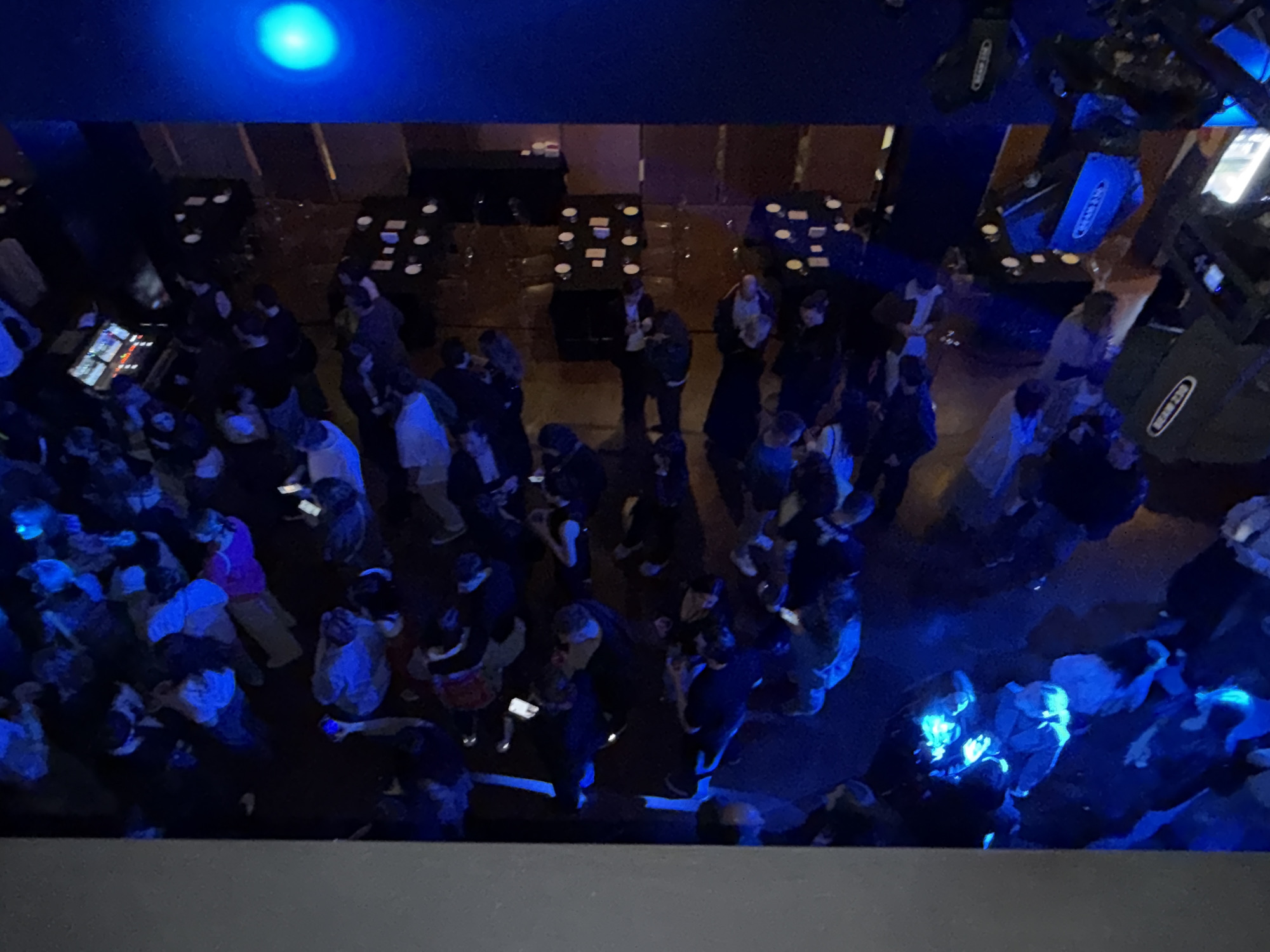 This overhead shot captures a bustling indoor social event or party in Tbilisi, Georgia, bathed in an intense blue artificial light. Numerous people are gathered throughout the venue, mostly standing and mingling. The dark surroundings, illuminated selectively by the vibrant blue light, create a nighttime atmosphere.
In the background, several tables are arranged, many appearing set with plates and glasses, indicating a dining area or event space where food might have been served. To the right, stage lighting equipment, specifically moving head lights, is visible. One piece of equipment clearly displays the text "BEAM 230", and other partially visible branding suggests "CHAUVET". On the left side, a control panel or screen with multiple illuminated buttons is visible, possibly for audio or lighting technicians.
The blue lighting casts a strong glow on individuals and surfaces, with some people's faces and clothing reflecting the light. Many attendees are seen holding and looking at their phones, which also emit a blue light, blending with the venue's illumination. The perspective from above provides a wide view of the energetic, crowded scene, suggesting a lively social gathering.