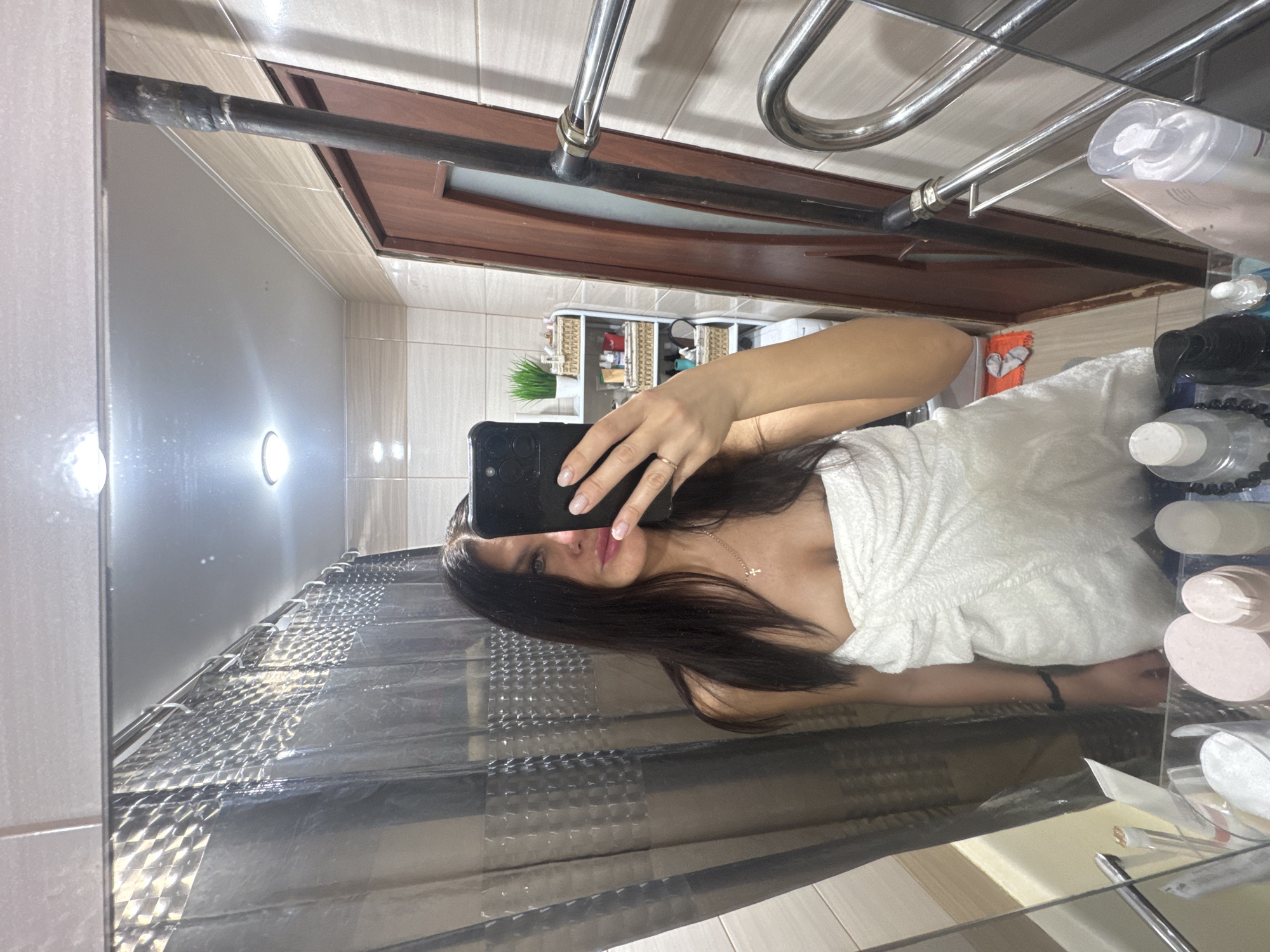 The provided image is a mirror selfie, rotated 90 degrees clockwise, depicting a woman in a bathroom. The woman, who has long, dark hair, is visible from the head to just above the waist. She is wrapped in a white towel or bathrobe, revealing her shoulders and a small cross necklace around her neck. She holds a black smartphone with her right hand to take the photo, and a ring is visible on her finger. Her expression is neutral as she looks towards the camera.
The reflection reveals a bathroom setting with light-colored, possibly cream or beige, tiled walls and floor. In the background, a brown wooden door or cabinet frame is visible, along with a chrome-plated metal pipe system, likely a heated towel rail, attached to the wall. To the left of the woman, a clear or translucent shower curtain with a textured or patterned surface hangs. Various bathroom essentials and toiletries, including bottles, containers, and cosmetic pads, are arranged on shelves or surfaces in the background and on the counter. Two circular light fixtures are mounted on the tiled wall. A small green artificial plant adds a touch of decor to a shelf. No discernible text is visible on any of the objects.