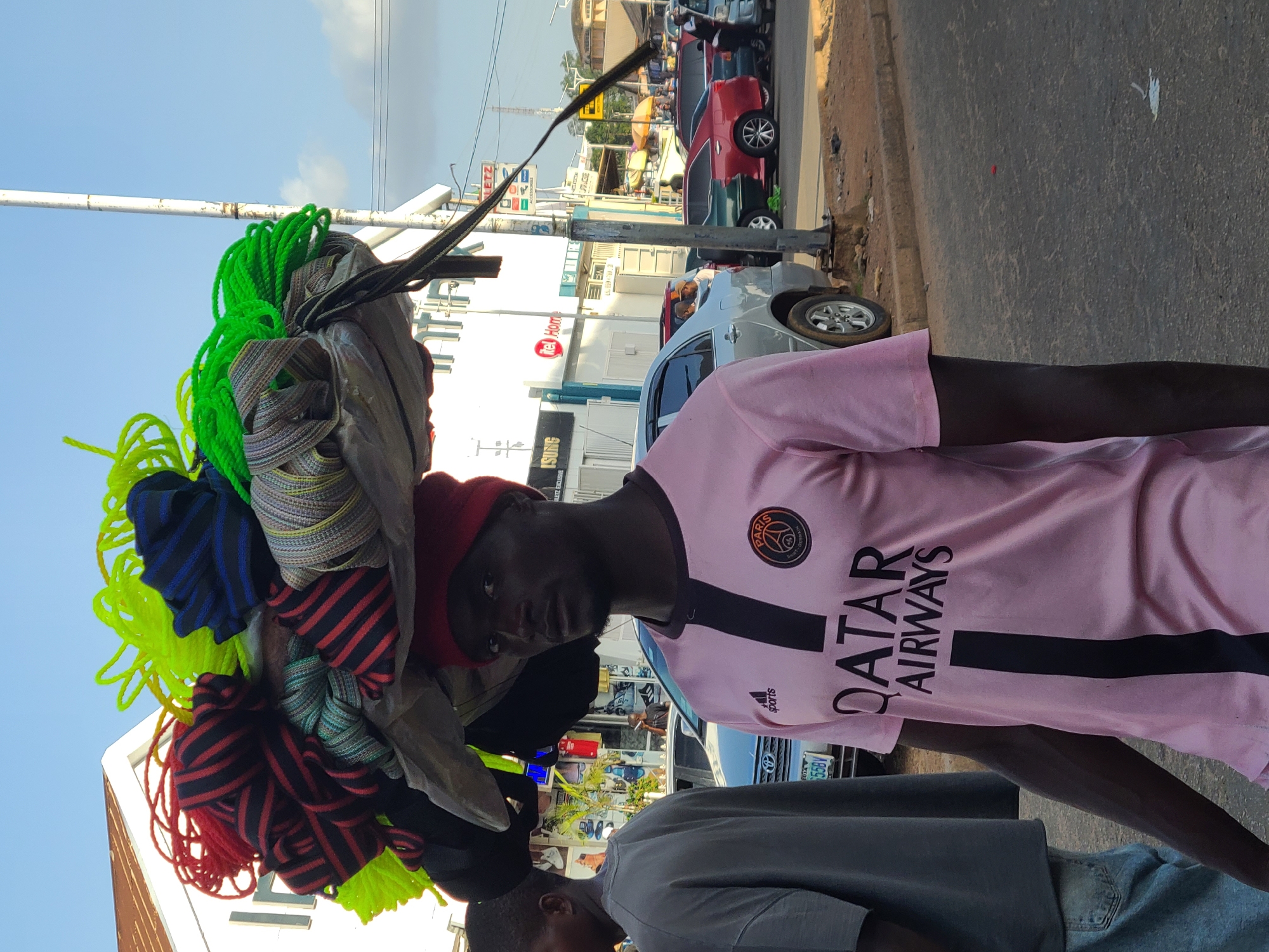 A Black man wearing a pink football jersey with "QATAR AIRWAYS" written on it and a red beanie is walking, carrying a large bundle of colorful ropes on his head. The ropes are coiled and appear to be neon green, red and black striped, and blue and black striped. He is walking on a paved street in Jos, Nigeria. Behind him, other people and vehicles are visible, including a silver car, a red car, and a green car. Buildings line the street, one of which has "Te Home" written on it. The sky is clear and blue.