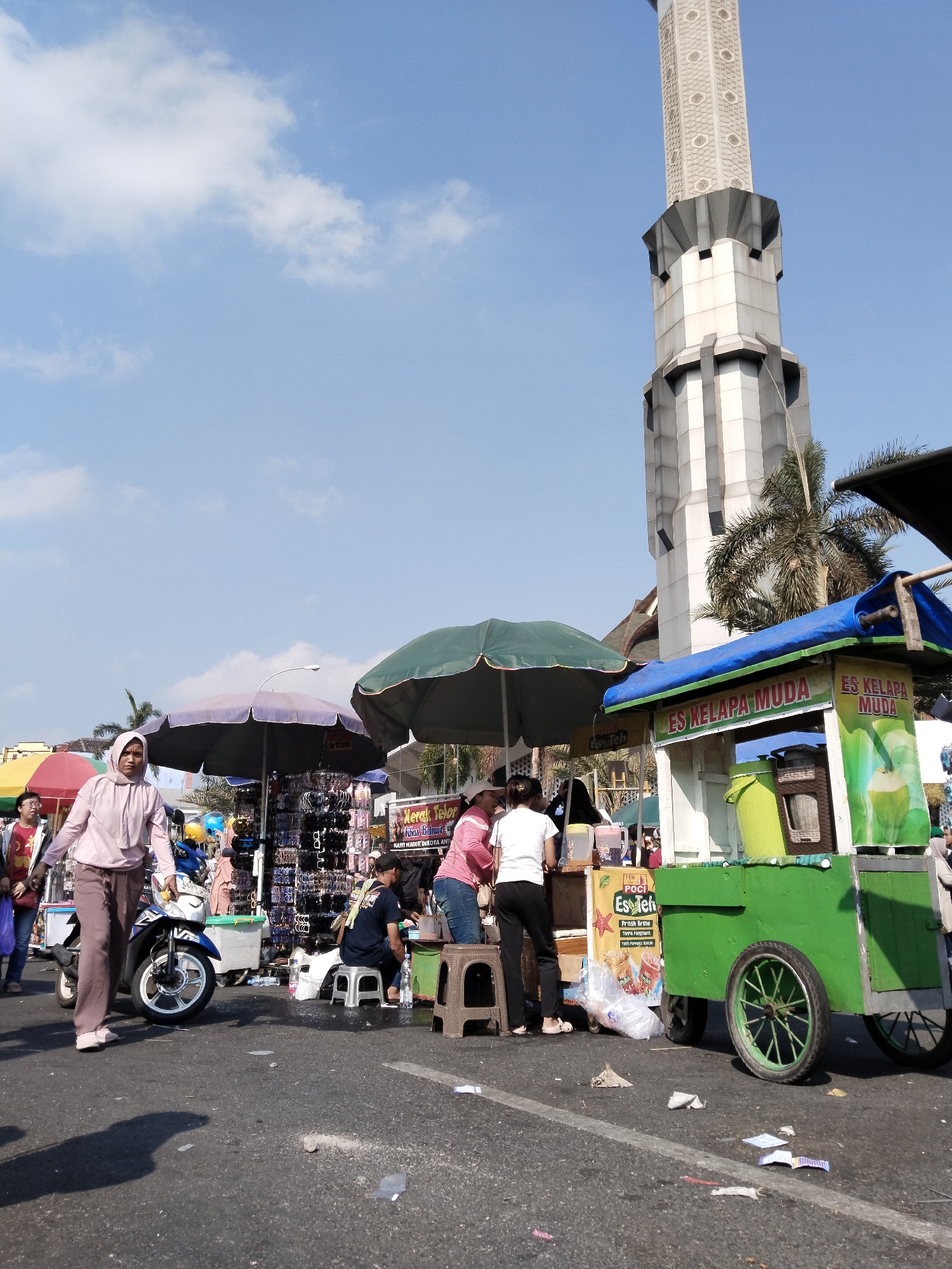 This is a street-level, full-shot photograph taken on a sunny day in Kecamatan Pati, Indonesia. The scene is set along a street where several vendors are operating. Dominating the background is a tall, modern minaret of a mosque, characterized by its grey, angular design and patterned upper sections. In the foreground and midground, there are multiple stalls and carts selling various items.
To the right, a bright green cart with a blue tarp roof displays signage for "ES KELAPA MUDA" (Young Coconut Ice), indicating it's a beverage stand. A large green bucket and a woven basket are visible on the cart. In front of the cart, two individuals, a woman in a pink shirt and another in a white shirt and black pants, are interacting with the vendor.
To the left of the green cart, there are several other stalls. One stall, covered by a dark green umbrella, appears to be selling accessories like sunglasses, displayed on racks. Another stall, under a light purple umbrella, also seems to be selling similar items. A woman in a light pink hooded jacket and brown pants is walking past these stalls, with a blue and white motorcycle beside her. She is looking towards the camera.
Further to the left, there are more vendors and people. A woman in a red shirt is visible near a yellow and red umbrella. The ground is paved asphalt, with scattered debris including papers and plastic wrappers. The sky is blue with scattered white clouds. The overall impression is of a busy outdoor market or street fair.