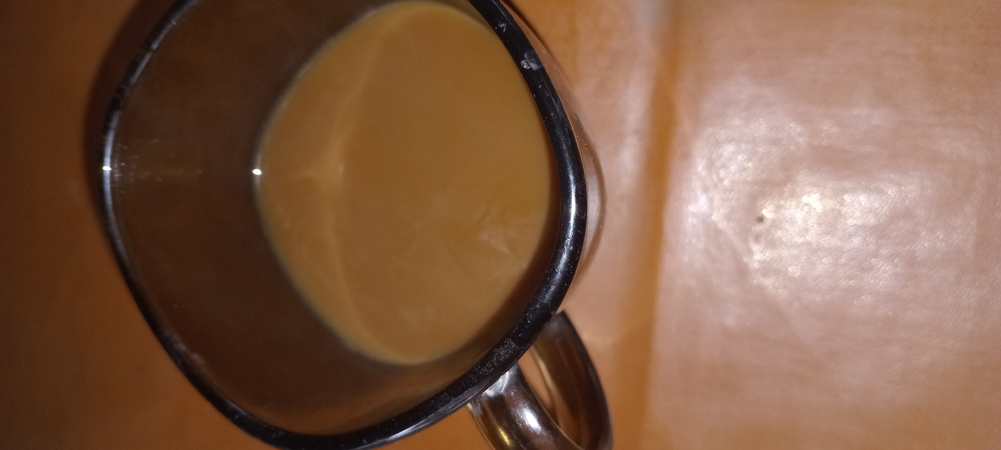 The image shows a close-up, high-angle view of a glass mug filled with a light brown liquid, likely coffee with milk or cream, resting on a wooden surface. The mug has a dark rim and a metallic handle. The background is a blurred, warm-toned surface, possibly a table or countertop. There are no people present in the image. The scene is indoors. The image does not contain any information to determine its location in the city of Koviahy, Ukraine, nor does it provide any details related to forensic analysis or an event.