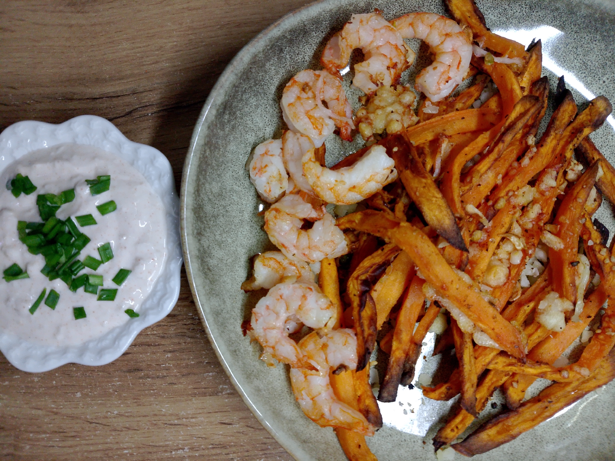 The image shows a plate of food consisting of shrimp and sweet potato fries, along with a small dish of dipping sauce topped with chopped chives, placed on a wooden surface. The location and city are not discernible from the image.