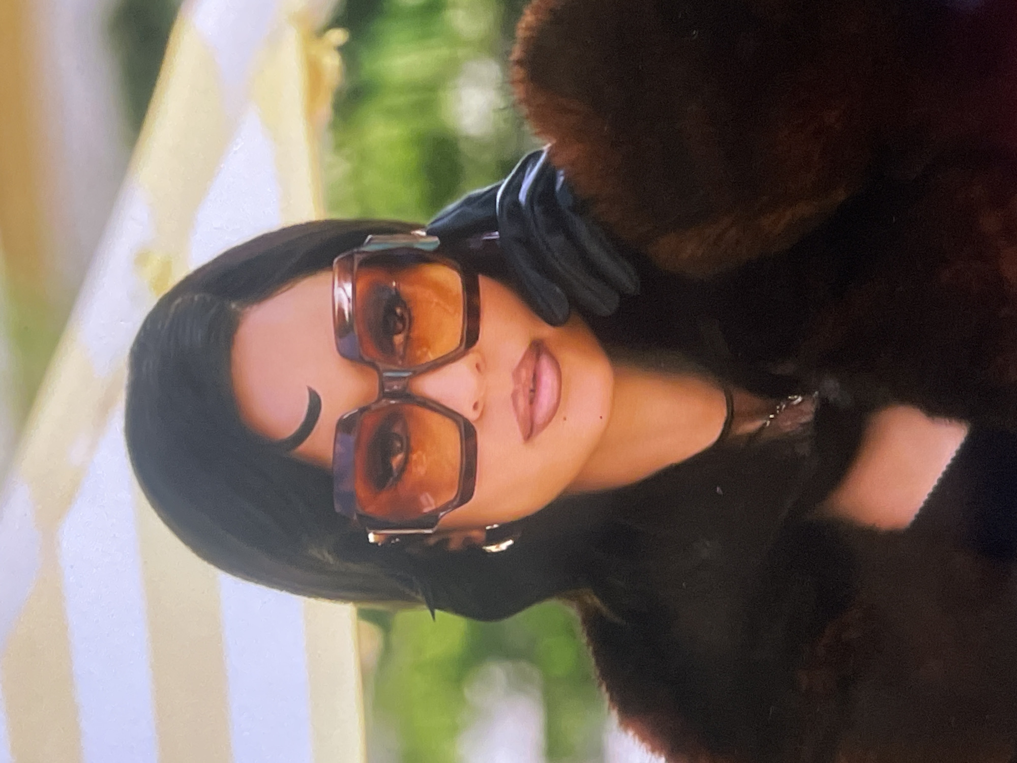 A close-up, eye-level portrait shot features a woman looking directly at the viewer, set outdoors in Poltava, Ukraine. She has dark hair and is wearing distinctive, large, geometric-shaped sunglasses with thick, dark frames and brown-tinted lenses. Her lips are slightly parted, showcasing a light pink or nude lipstick.
She is dressed in a dark, possibly brown, fur or faux-fur coat, with a gloved hand (dark, possibly leather or fabric) resting near her temple, touching her hair or sunglasses. A thin dark necklace is visible around her neck.
The background is softly blurred, showing a green area indicative of foliage or grass on the right side of the frame (when oriented correctly). To the left, a light-colored structure with vertical stripes of white and pale yellow is visible, suggesting an awning, tent, or decorative wall. The overall lighting is bright and natural, indicating a daytime setting. The attire, particularly the fur coat and gloves, suggests cooler weather. There is no visible text in the image.