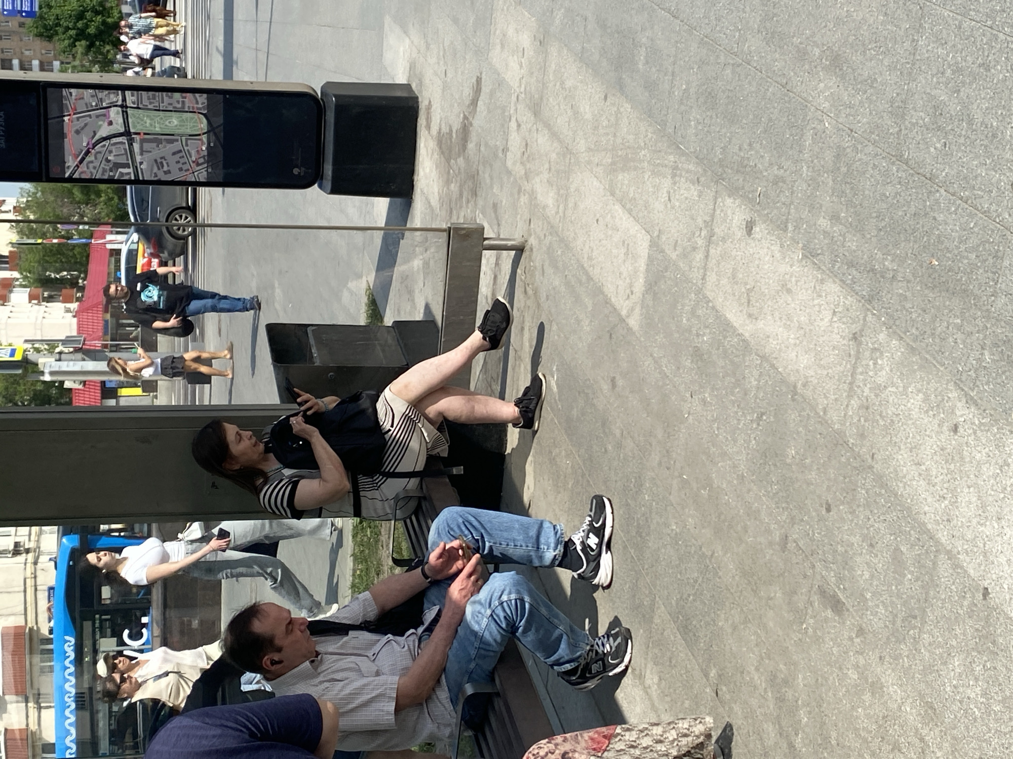 The image captures a scene in Moscow, Russia, near what appears to be a public transit stop or information point. There are several people visible. A woman is seated on a bench with her legs stretched out. Nearby, a man is also sitting. Another person, wearing a blue shirt, sits closer to the viewer. A digital display shows a map of Moscow. Reflections in the glass surface of the information point show other people, buildings, and a bus. The environment consists of a paved area, buildings, and trees in the background.