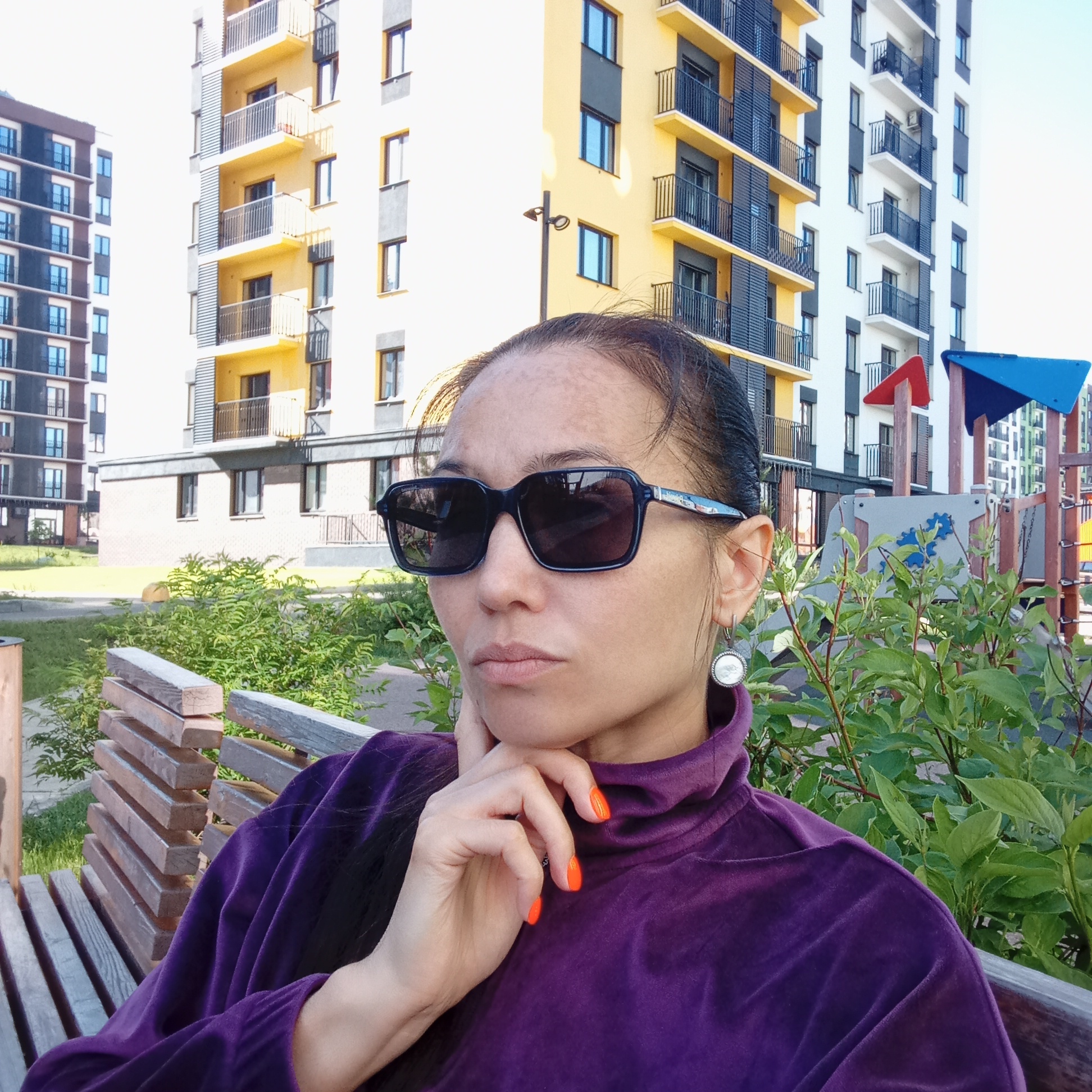 A mid-shot captures a woman seated on a wooden bench outdoors on a sunny day. She is looking thoughtfully towards her left, with her right hand resting on her chin. She wears dark sunglasses and a dark purple, possibly velvet, high-neck top. Her fingernails are painted a vibrant orange. A white, round earring with a textured silver border is visible on her left ear.
In the background, several modern multi-story residential buildings are visible under clear skies. One prominent building features bright yellow and white facades with dark gray or black balconies and window frames. Another building, partially visible to the far left, has a darker color scheme, possibly brown and dark gray. Green foliage, including bushes, is scattered around the scene, with some obscuring the lower part of the background. A colorful children's playground with red and blue elements is visible behind the woman and the bushes. The bright lighting suggests it is daytime with good weather. No discernible text is visible in the image.