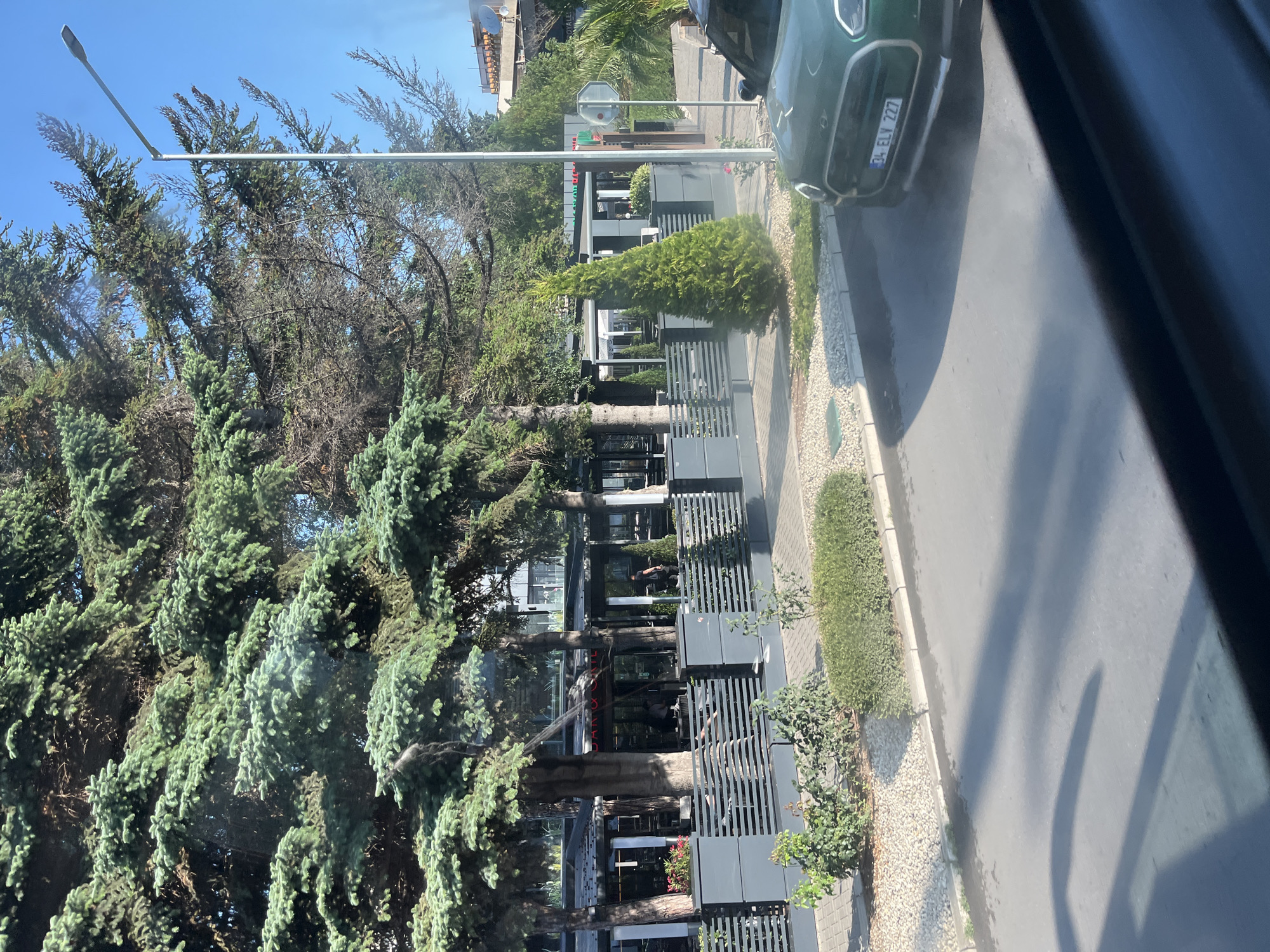 The image depicts a street scene in Nesebar, Bulgaria, with a focus on a restaurant or cafe area. Large coniferous trees line the sidewalk, providing shade. The establishment has an open-air seating area with tables and chairs, partially enclosed by a modern gray fence. A green car with a Turkish license plate (34 ELV 227) is parked on the right side of the street, partially in the frame. A lamppost stands near the trees, and the sky is clear and blue. Shadows from the trees and the car are cast on the asphalt. The overall impression is a sunny day in a coastal town, possibly a resort area.