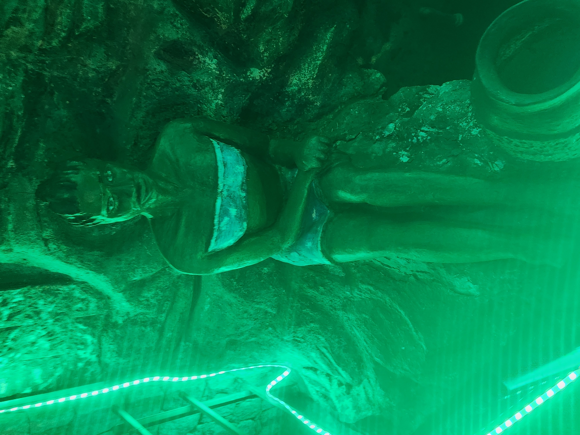The image depicts a statue of a woman wearing a swimsuit, lying on a rocky surface inside a grotto. There is a large vessel above the woman. The scene is lit with green and white LED strip lights. This location is in Jos, Nigeria.