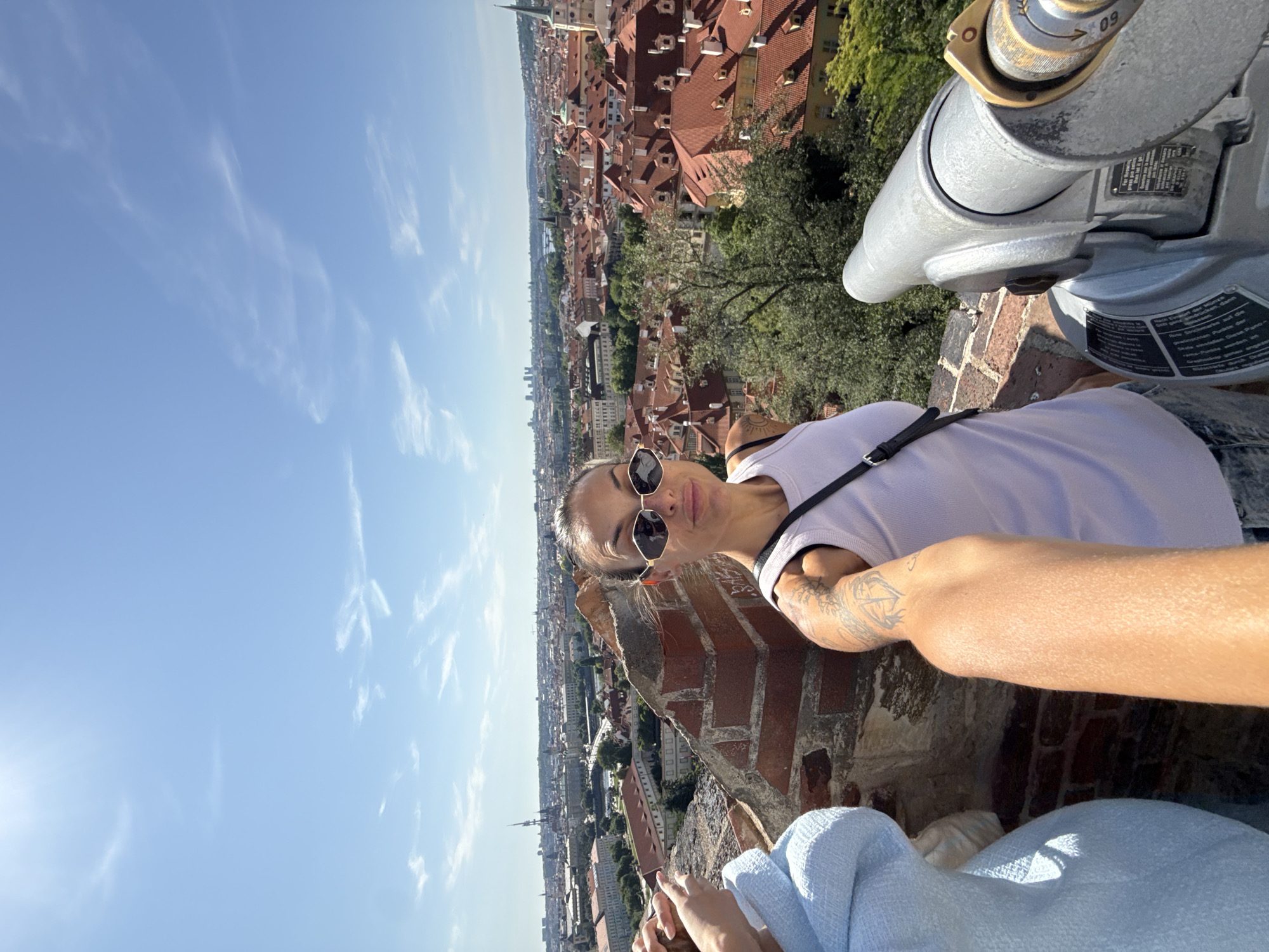 A woman with sunglasses and tattoos takes a selfie while standing on a brick wall overlooking a city with red-tiled roofs, likely Prague, Czechia. She is wearing a white tank top and denim shorts. A coin-operated telescope is visible in the foreground. The sky is blue with scattered clouds.