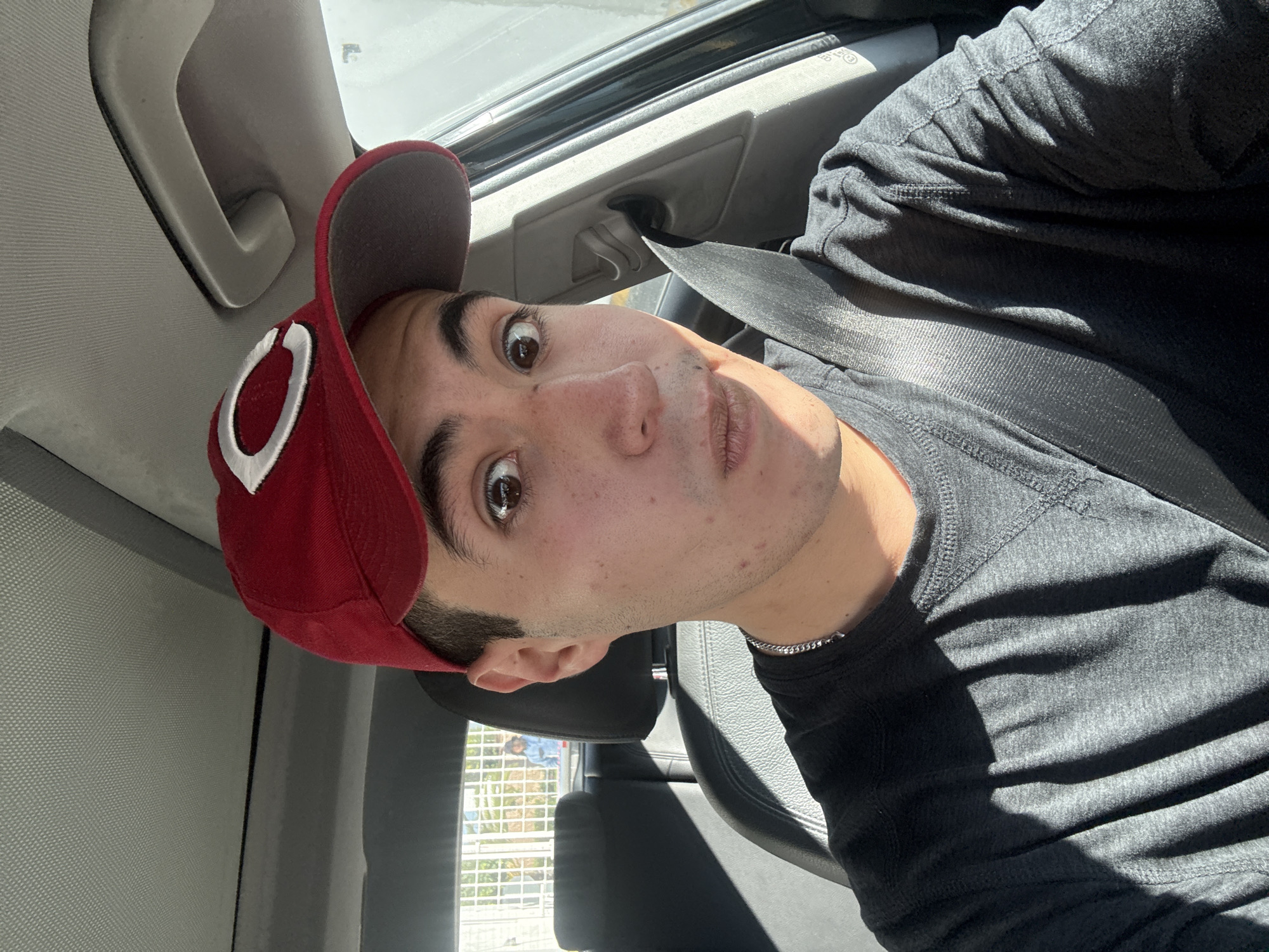 A young man with dark hair and brown eyes, wearing a red baseball cap with a white "C" logo and a dark gray long-sleeved shirt, is taking a selfie in the driver's seat of a car. The man has a surprised or playful expression, with his mouth slightly open and his eyebrows raised. A silver chain is visible around his neck. The car's interior is visible, including the black leather seat, a gray seatbelt across his chest, and part of the car's ceiling. Sunlight casts shadows on his face and shirt. In the background, through the car window and the rear window, a person is visible near a fence outside the car. The image appears to have been taken in San Bernardino Tlaxcalancingo, Mexico, indicated by context provided separately from the image itself.