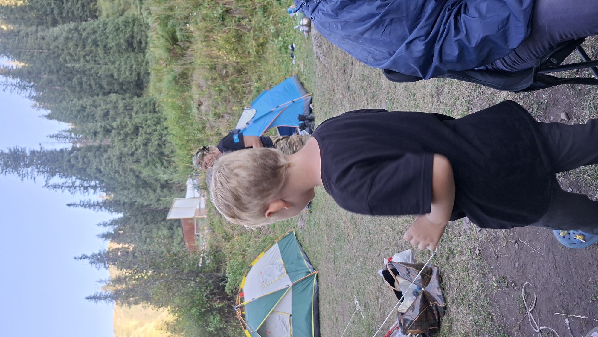 A young boy with blonde hair and a black t-shirt is standing in the foreground, facing away from the camera. To his left, a green and yellow tent is set up on grassy ground. In the background, a person wearing a bandana and camouflage pants stands near a blue tent. Behind the tents, a dense forest of pine trees covers a hillside. The sky is clear and blue. The scene appears to be a campsite in a mountainous or forested area. The image does not provide any specific details about the city of Batan, Kazakhstan.