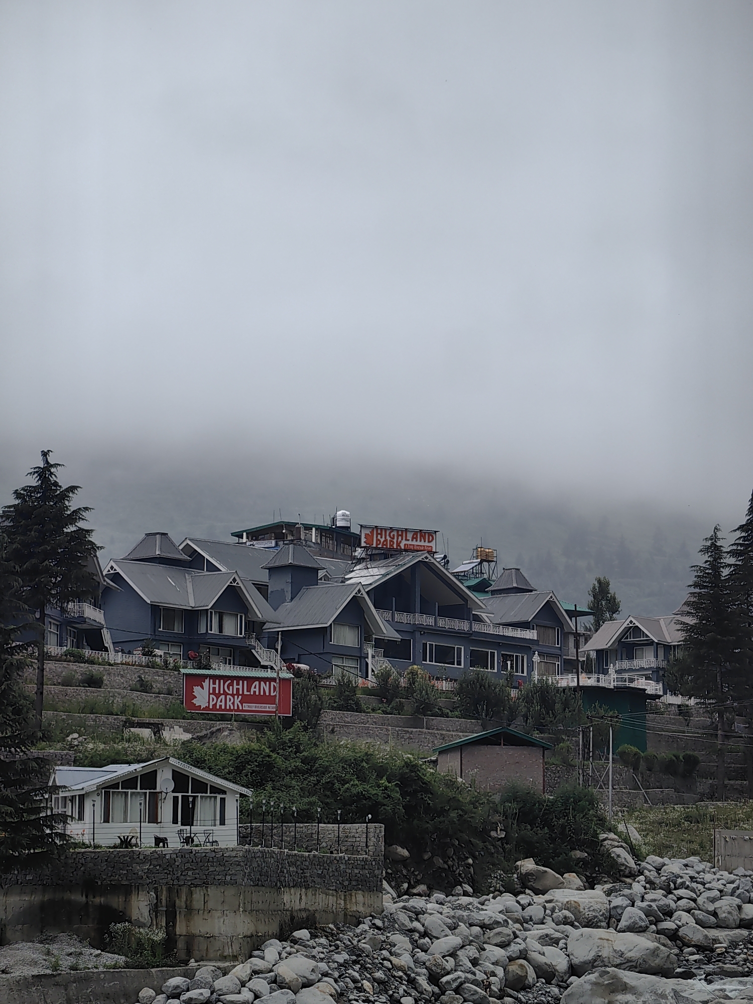 In Barheta, India, a cluster of blue buildings with gray roofs is situated on a hillside. The buildings appear to be part of a resort or hotel complex. A large red sign with white lettering reading "HIGHLAND PARK" is prominently displayed on the hillside, below the main buildings. A smaller white building with a pitched roof is located at the bottom of the frame, closer to the viewer, and appears to be a separate structure within the complex. The foreground is filled with a rocky riverbed, suggesting the resort is located near a water source. Tall pine trees are visible on the left and right sides of the image. The sky is overcast and foggy, with the fog obscuring the background hills and mountains. No people are visible in the image.